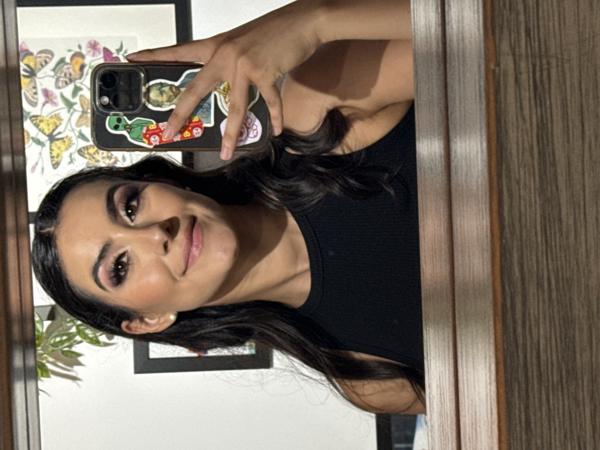 A medium shot captures a woman, likely in her early to mid-twenties, taking a selfie in a mirror. She has dark hair, styled with waves, and is wearing a black top. Her makeup includes a prominent eyeshadow, and a pearl earring is visible.
She holds a smartphone with stickers on the back. The mirror reflects part of a room interior, including a framed picture with butterfly illustrations and a small plant.
The location is not discernible to Puebla, Mexico.