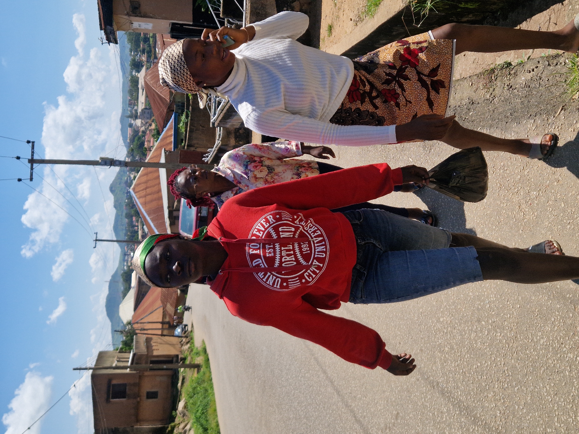 Three women walk along a paved road in Kabong, Nigeria, under a bright, partly cloudy sky. The woman in the foreground on the left wears a red hooded sweatshirt and denim shorts, carrying a black plastic bag. To her right, a woman in a white turtleneck and a patterned skirt talks on a mobile phone. Behind them, a third woman with red hair glasses and a floral shirt walks also. The road is lined with buildings and vegetation, and in the distance, hills are visible. A utility pole with wires stands on the right side of the road.