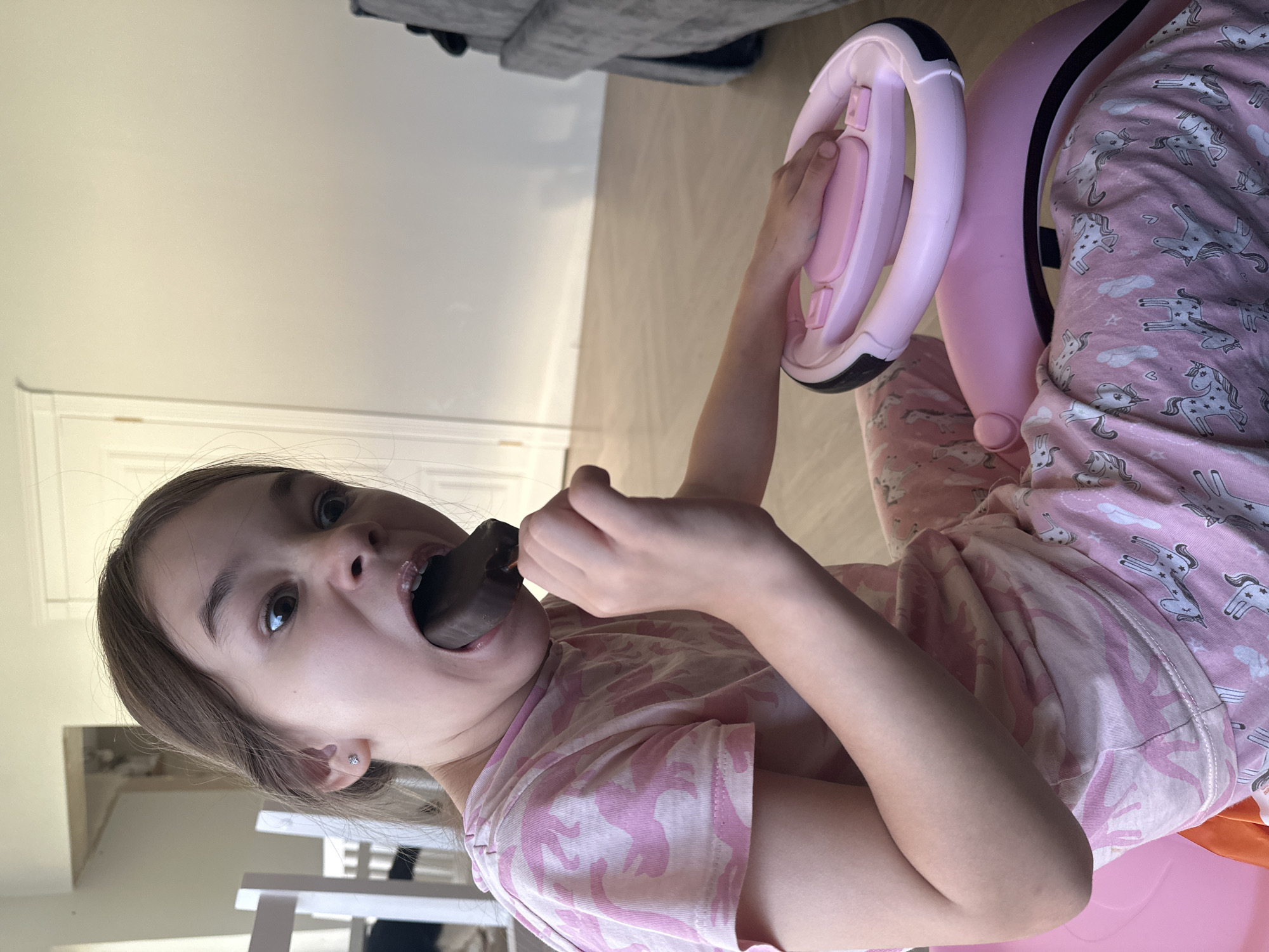 A child, likely female, is indoors, apparently seated on a pink ride-on toy with a pink steering wheel. She is wearing pink pajamas patterned with unicorns. She is eating a dark-colored treat, possibly chocolate, with a portion of it in her mouth. The setting is a room with light-colored walls and flooring. Furniture is partially visible. The location is unknown and there is no evidence it is Kharnavy, Russia.