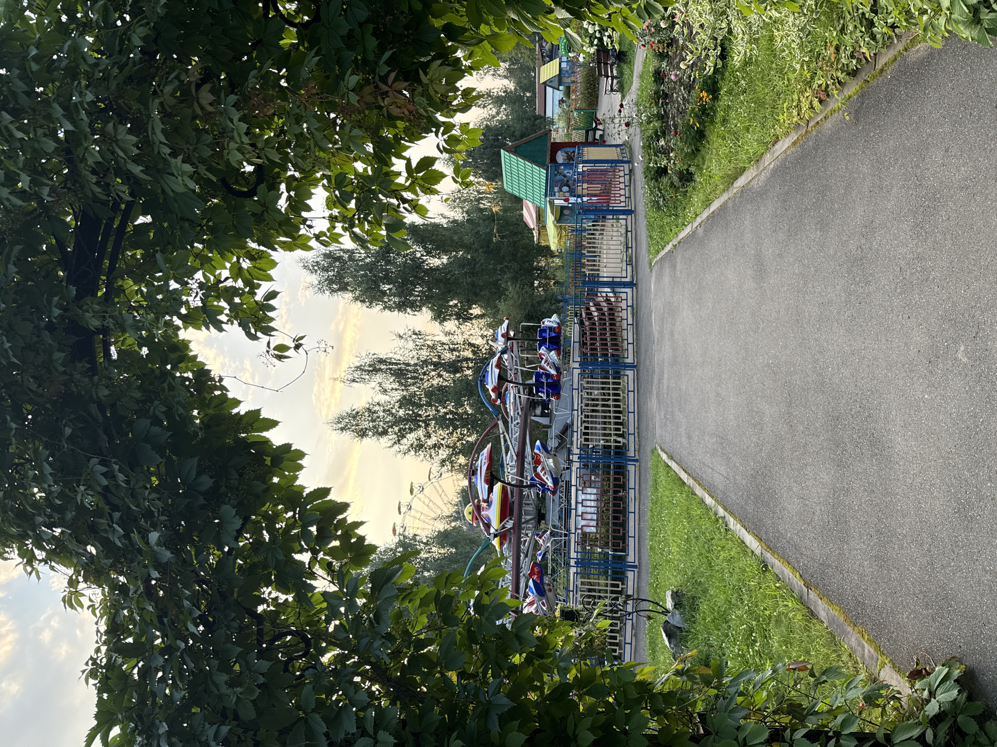 This image depicts an amusement park in the city of Chaikovsky, Russia, framed by lush green foliage. A paved pathway leads towards a children's amusement ride, which features colorful, airplane-shaped seats. The ride is enclosed by a blue and red fence. In the background, a Ferris wheel can be seen, along with a few small buildings with green roofs. The sky above is partly cloudy with the soft light of either sunrise or sunset. The area appears to be a public park, with grass, flower beds, and benches visible. No people are present in the image.