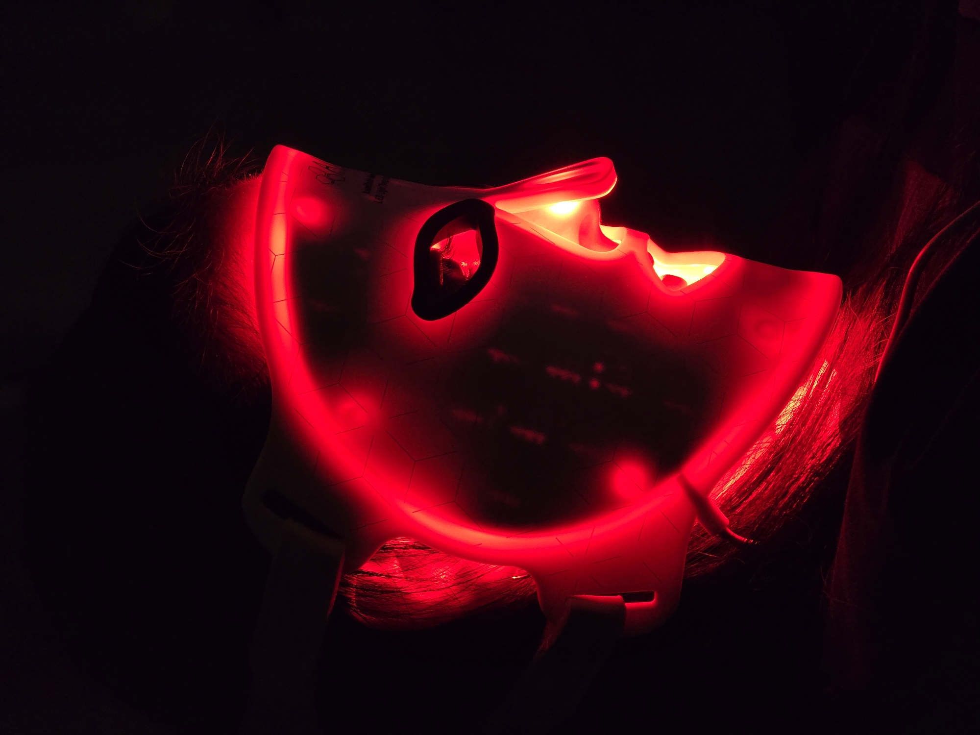 The image captures a close-up of a person wearing a white LED light therapy face mask that is actively glowing with a vibrant red light. The mask covers the entire face, with distinct cutouts for the eyes and nose, and an area for the mouth. Its surface features a subtle hexagonal grid pattern through which the red light emanates. The intense red light illuminates the edges of the mask and brightly highlights the person's hair, which is visible around the mask's perimeter and beneath it, appearing reddish-orange due to the glow.
The scene is set against an almost completely dark background, which dramatically accentuates the glowing mask and the light it casts. This darkness suggests the photograph was taken in a dimly lit room or at night, creating an intimate and focused atmosphere for the activity. The primary activity depicted is the use of an LED light therapy device, likely for skincare or cosmetic purposes.
Notable details include faint, partially obscured text visible on the upper left side of the mask, where words such as "POWER" and "LONGPRESS" can be discerned, along with some small symbols, indicating control functions or branding. The person's facial expressions are not visible due to the mask. The overall impression is of a modern beauty or wellness treatment being performed in a private setting.