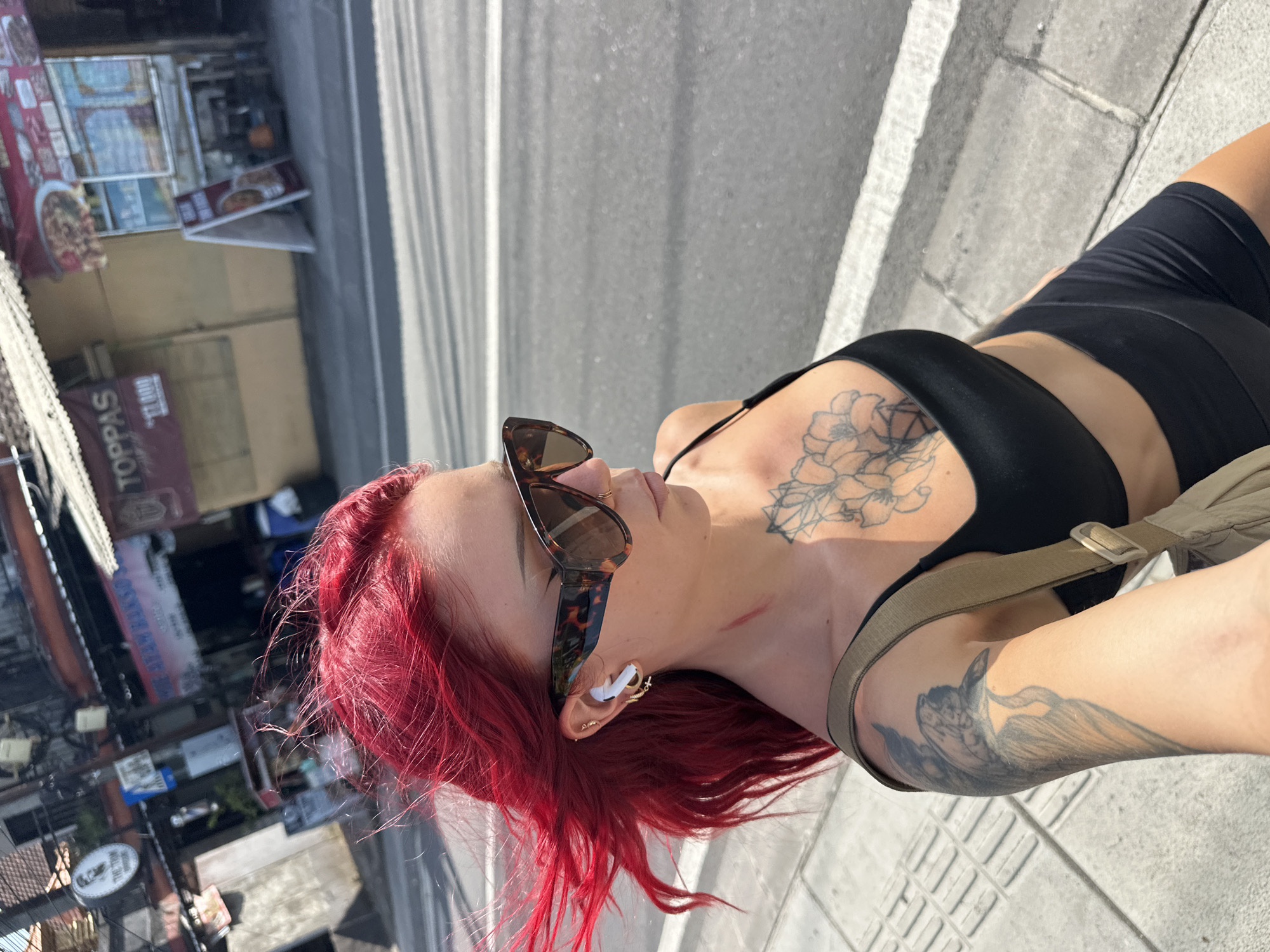 A woman with bright red hair is standing outside in North Kuta, Indonesia, possibly on a sidewalk, next to a street. She is wearing sunglasses and an earbud in her ear. She is also wearing a black sports bra and black shorts. She has tattoos on her chest and arm. A building with signage (one reads "Toppas") is visible in the background.