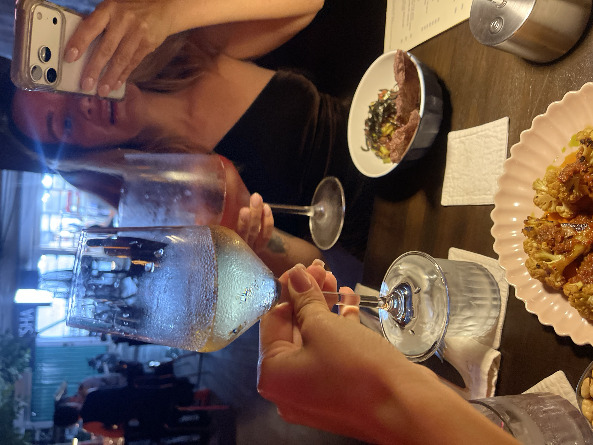 In the city of Mengwi, Indonesia, at a dimly lit restaurant, two women are seated at a table. One woman, in the foreground, holds a glass of white wine. The other woman, in the background, is taking a selfie with her phone while holding a glass of rosé. The table is set with food, including a dish of cauliflower and what appears to be a small appetizer in a bowl. The ambiance suggests a casual dining experience.