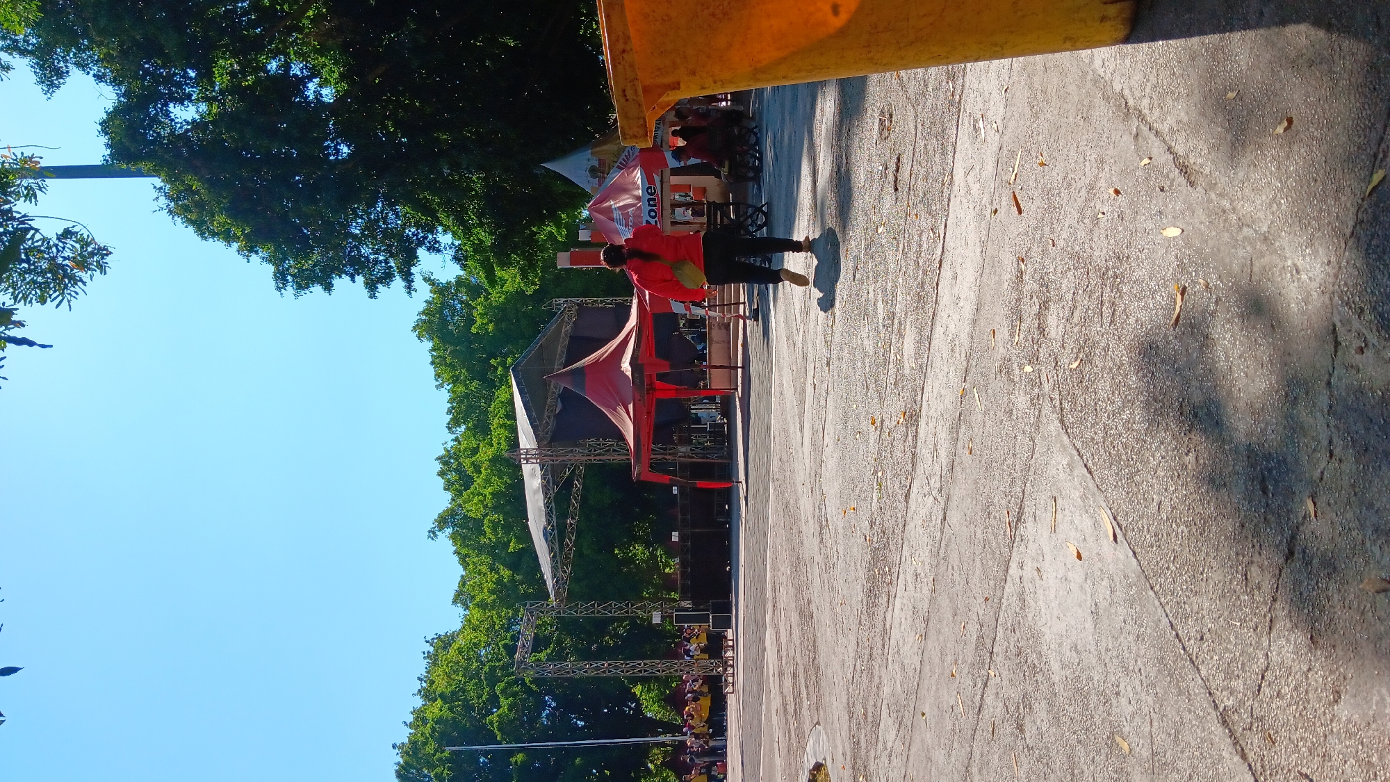 In a sunny outdoor setting, likely a public park or plaza in Nganjuk, Indonesia, a person is walking away from the camera in the foreground. They are wearing a red jacket and dark pants, with a light-colored bag slung across their body. The ground is paved with concrete, with some fallen leaves scattered across it. In the middle ground, a stage setup is visible, featuring a large black tent or canopy with a metal truss structure. In front of the stage, there's a smaller red tent. Several people are standing in groups, some near the stage and others behind tents, possibly attending or working at an event. Further in the background, lush green trees form a natural backdrop. To the right of the frame, a portion of a large yellow trash bin is visible, partially obscuring the scene. The sky above is a clear, bright blue.