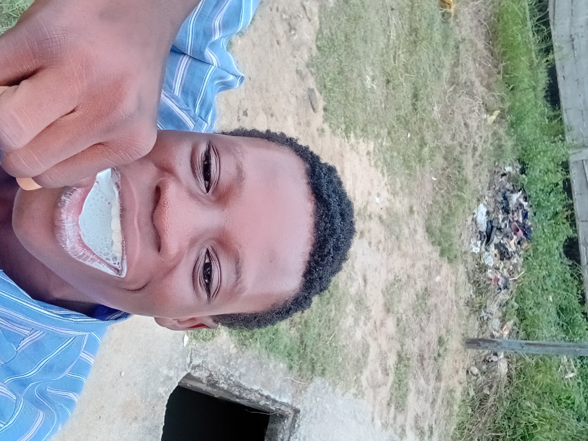 A close-up, selfie-style, low-angle shot captures a young Black individual from the chest up, outdoors. The individual is wearing a blue and white striped collared shirt. Their mouth is open, revealing teeth with what appears to be toothpaste or foam. A hand is visible on the left side of the frame, holding a toothbrush near the individual's mouth. The background shows a mix of dry, patchy grass, dirt, and a pile of refuse. A concrete structure with an opening is present on the right side of the frame, and a wooden structure or fence is visible in the distance. The lighting suggests it is daytime.