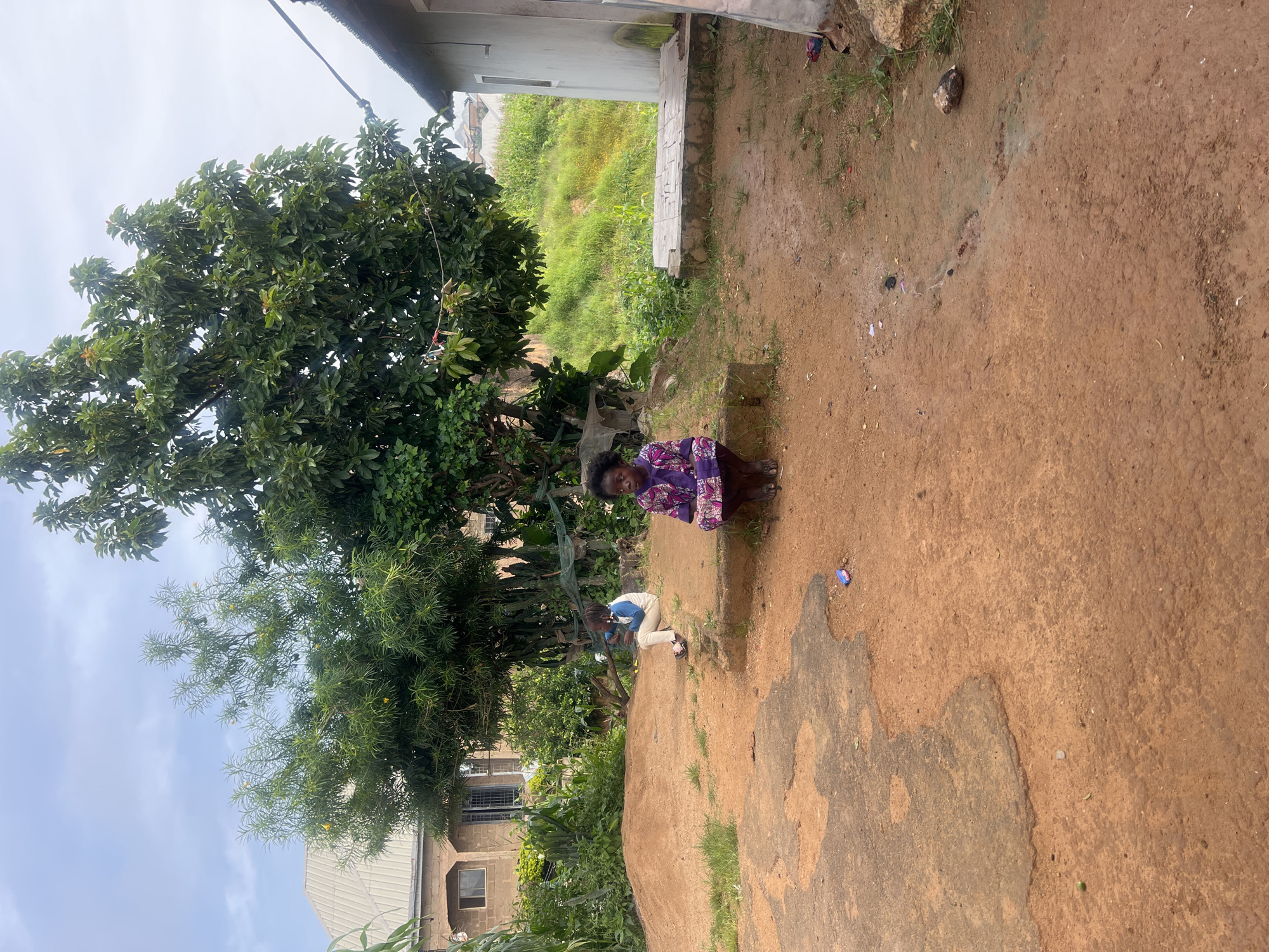 A wide shot captures an outdoor scene with two young children on a dirt ground in front of a building. The child on the right, a girl, is crouched down, wearing a purple and white patterned garment. To her left and slightly behind her, another child, possibly a boy, is seated on a raised edge, dressed in a white and blue shirt and light-colored pants. They are in what appears to be a residential area, with a building that has a metal roof visible in the background. Lush green trees and vegetation surround the area, and a patch of uneven, possibly repaired, asphalt is in the foreground. The ground is predominantly reddish-brown dirt, with some sparse green grass. The sky is overcast. This scene is located in Jos, Nigeria, a city known for its elevated plateau and temperate climate.