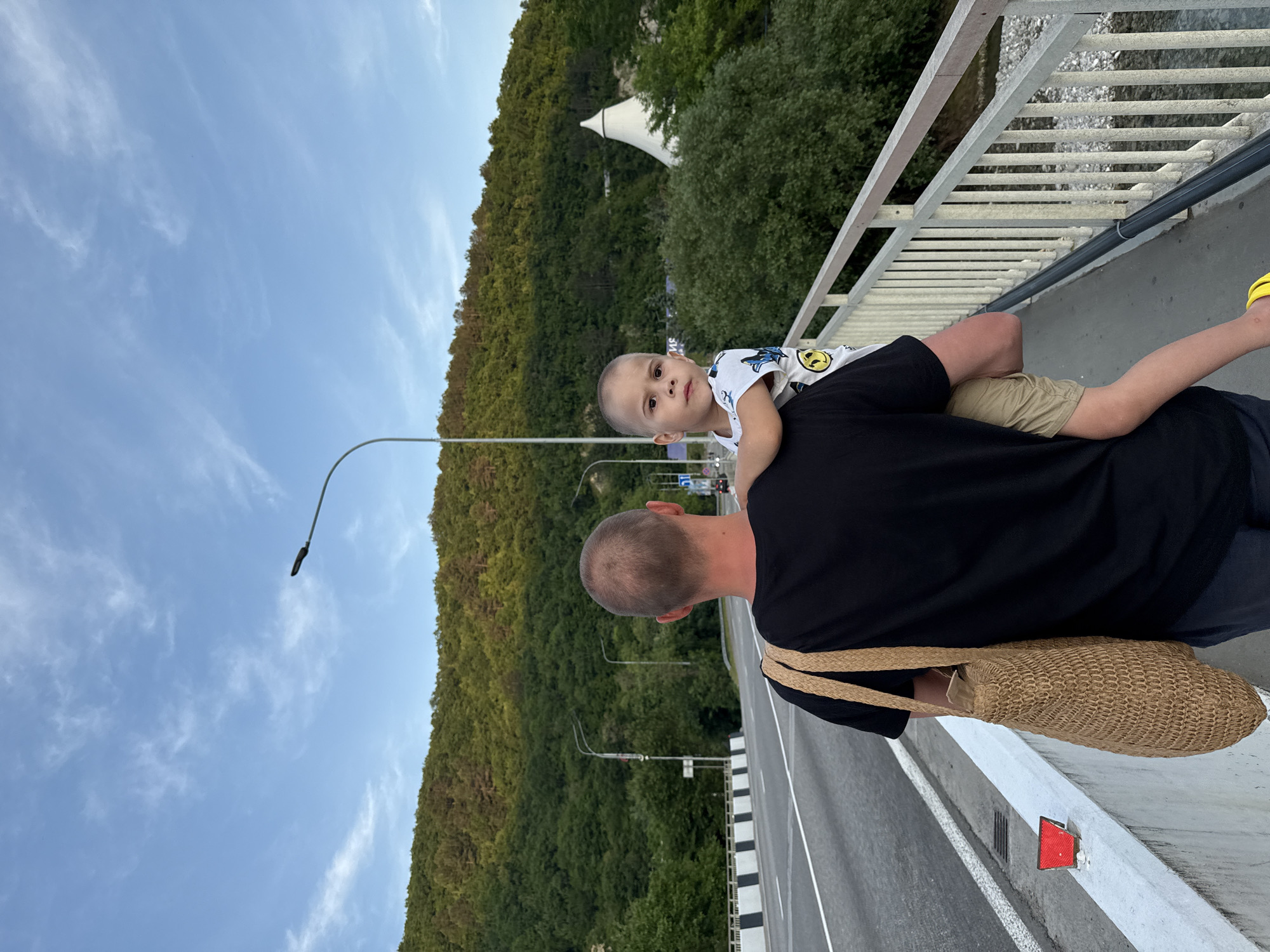 The image depicts an adult male carrying a young child on his left hip, walking on the pedestrian side of a bridge or elevated road in Nebug, Krasnodar Krai, Russia. The man, viewed from the back, has short, light-colored hair and wears a black t-shirt and dark trousers. A woven tan tote bag is slung over his right shoulder. The child, a boy estimated to be 3-5 years old, faces the camera with a neutral expression. He wears a white t-shirt featuring a smiley-face design and khaki shorts; a yellow band is visible on his left wrist.
To the left of the subjects, a white concrete road barrier with black and white stripes separates the pedestrian path from a multi-lane paved road, marked with white lines. A red rectangular reflector is affixed to the top of this barrier. A light grey metal guardrail with vertical posts runs along the right edge of the pedestrian path, overlooking a lower area, possibly a riverbed. Several streetlights on tall poles are visible, with one prominent black streetlight fixture extending from the right side of the frame towards the upper center.
The background features a densely forested mountain range under a light blue sky with scattered wispy white clouds. Against this backdrop, prominently positioned in the mid-ground on the right, stands a distinctive white conical building, identifiable as the Dolphinarium Aqua World in Nebug. Additional distant road infrastructure and buildings are partially visible among the trees on the left side of the mountain slope.