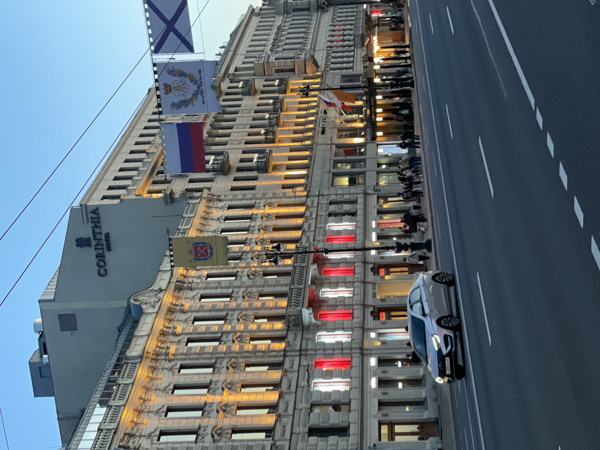 The image captures an evening or dusk scene on a wide urban street in Saint Petersburg, Russia. On the left, a large, ornate building with classical architectural details features numerous windows, many of which are illuminated with warm, golden light. Some lower windows display distinct red and white lights. Adjacent to this building, the upper facade of another building is visible, bearing the prominent text "CORINTHIA HOTEL".
Several flags are displayed above the street. From top to bottom, these include the Andreevsky flag (a blue saltire on a white field, the naval ensign of Russia), a flag featuring a golden crown and an elaborate crest, and the national flag of Russia (horizontal white, blue, and red stripes). Another smaller Russian flag and a flag with a red cross are also visible lower down.
On the right, a multi-lane asphalt road with clear white lane markings stretches into the distance. A light-colored car, with its headlights on, is driving in the foreground. Along the sidewalk to the left of the road, numerous pedestrians are visible, some gathered in small groups, suggesting a bustling evening atmosphere. The sky above is a clear, light blue, indicating the transition from day to night. Streetlights and power lines are also part of the urban landscape.
