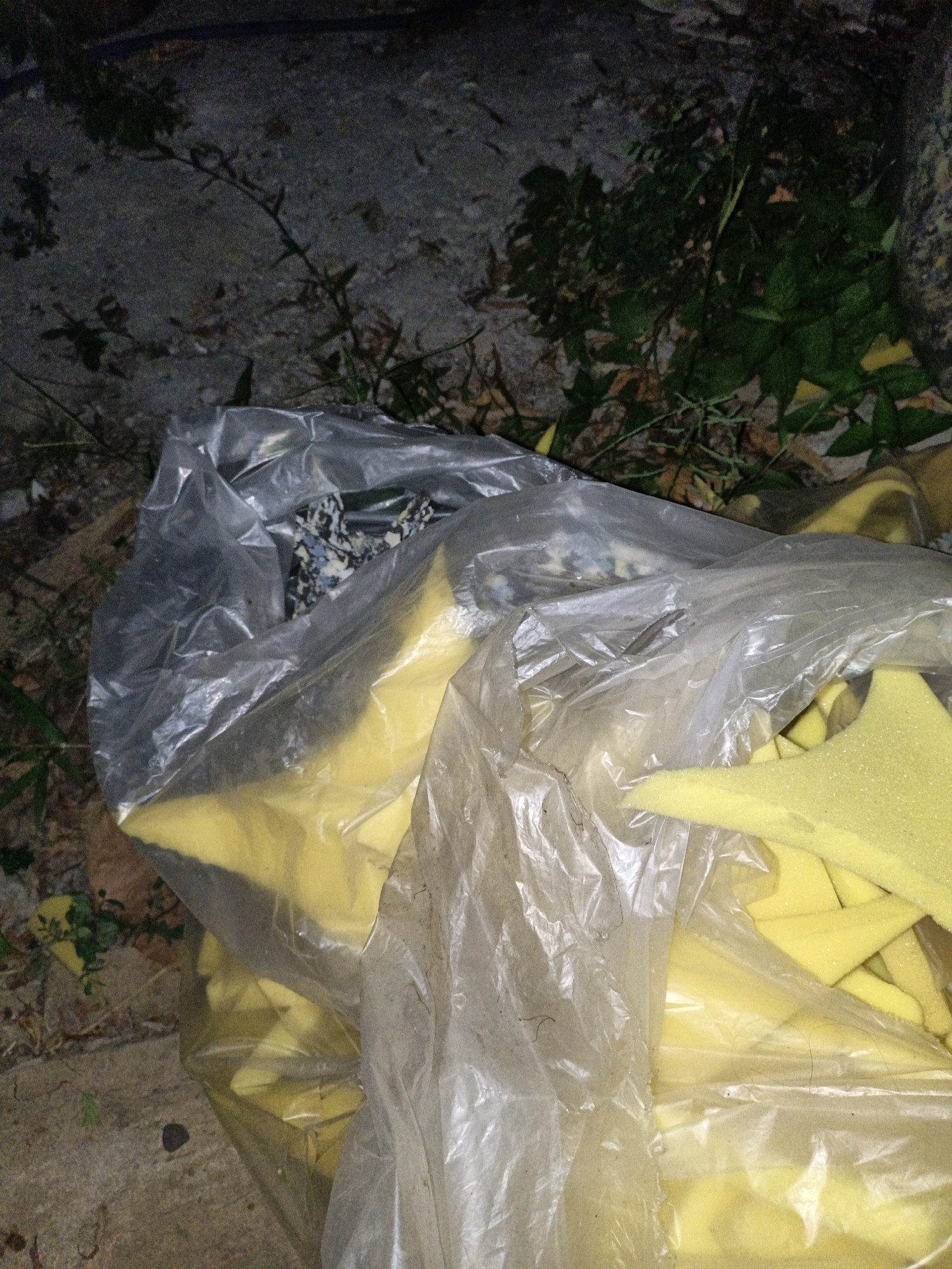 The image shows a clear plastic bag filled with yellow foam pieces. The bag is lying on a gritty, textured surface, possibly a road or a sidewalk, with some sparse green vegetation and dry leaves scattered around. In the background, there is a dark, rough wall-like structure and more vegetation. The lighting suggests it is either dusk or night. The location is identified as Cikal, Indonesia. The scene appears to be an outdoor setting, possibly a roadside disposal area.