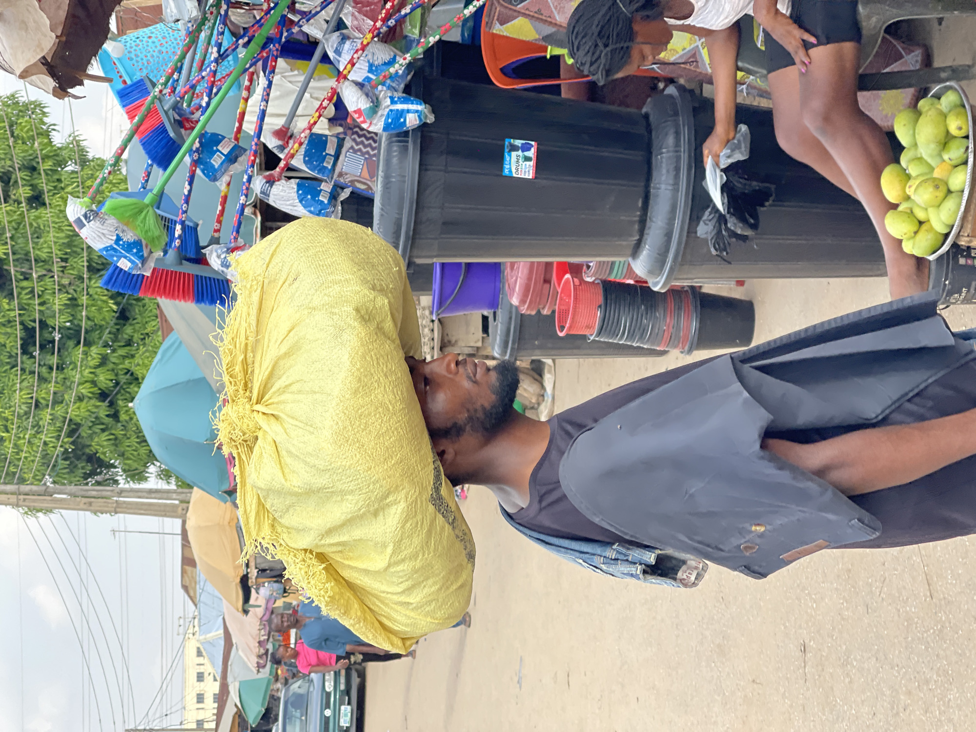 This photograph captures a bustling street market scene in Kuchingoro, Nigeria, under what appears to be a bright, possibly overcast, daytime sky.
In the foreground, a man walks, expertly balancing a large, textured yellow sack on his head. He has a dark beard and wears a dark t-shirt with a grey or dark blue vest over it, and a denim jacket tied around his waist. His gaze is directed forward and slightly upwards.
To the man's right, a market stall is visible, laden with various household goods. Hanging prominently are numerous colorful brooms with plastic bristles in red, blue, and green, and patterned handles. Below these, stacks of black plastic drums and buckets are arranged. One large black drum has a blue and white sticker with the word "DRUMS" clearly visible, along with smaller graphics. Other plastic items like red and purple buckets and baskets are also on display.
A woman is seated next to this stall, her legs visible. She is wearing a white top and black shorts, and her hair is styled in braids. She appears to be handling something black, possibly a plastic bag, near the drums. Beside her, a metal basin is filled with numerous green mangoes, indicating fresh produce is also available.
Further down the street, the scene suggests more market activity with blue and beige umbrellas providing shade over other stalls or vendors. Utility poles with wires crisscross the sky, and blurred figures of other people and a vehicle can be seen in the distance, underscoring the busy nature of the location. The ground is a light-colored paved or dirt surface.