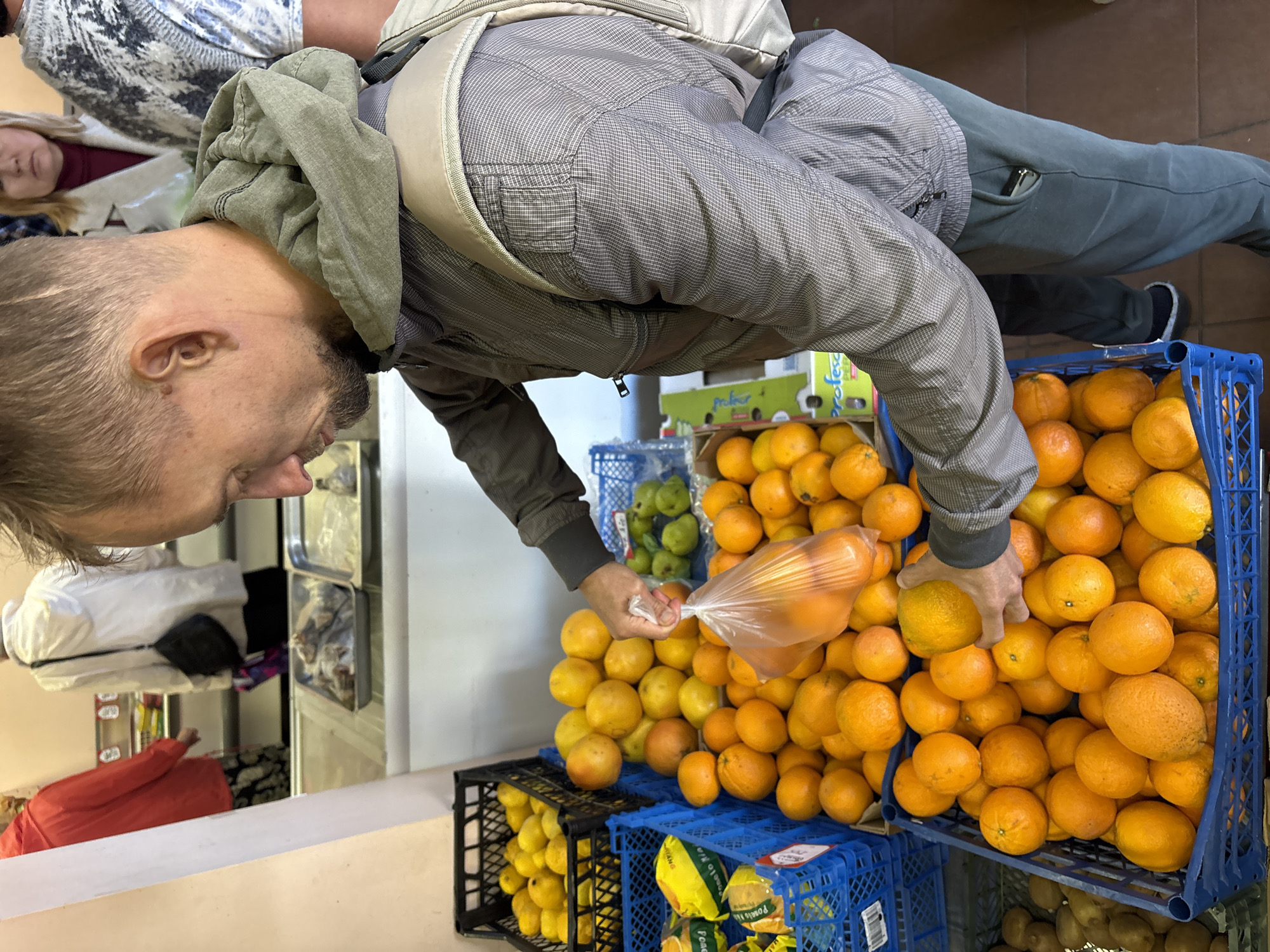 An indoor market scene in Gelendzhik, Russia, shows a man selecting oranges from a large display. The man, with short light brown hair and a beard, is wearing a grey hooded jacket over a light-colored patterned shirt and dark trousers. He is focused on bagging oranges, holding a clear plastic bag already containing several oranges with one hand and picking another orange from a blue plastic crate with his other hand.
The market stall is filled with numerous plastic crates, predominantly blue and black, overflowing with fresh produce. Large quantities of vibrant orange citrus fruits are prominently displayed. Other visible fruits include lemons in a black crate and green fruits, possibly limes or small apples, in another crate in the background. At the bottom right, a brown crate holds what appears to be kiwi fruits or potatoes.
In the background, two other individuals are partially visible. One woman with blonde hair, wearing a red top and a white cardigan, is seen looking downwards. Another person, mostly obscured, wears a blue and white patterned top. The setting features light-colored walls and a tiled floor. Visible text includes "pro.food" and "Profesor" on a green cardboard box, and "Pomelo" on some yellow packaging. The activity suggests a typical shopping experience at a local fruit market.