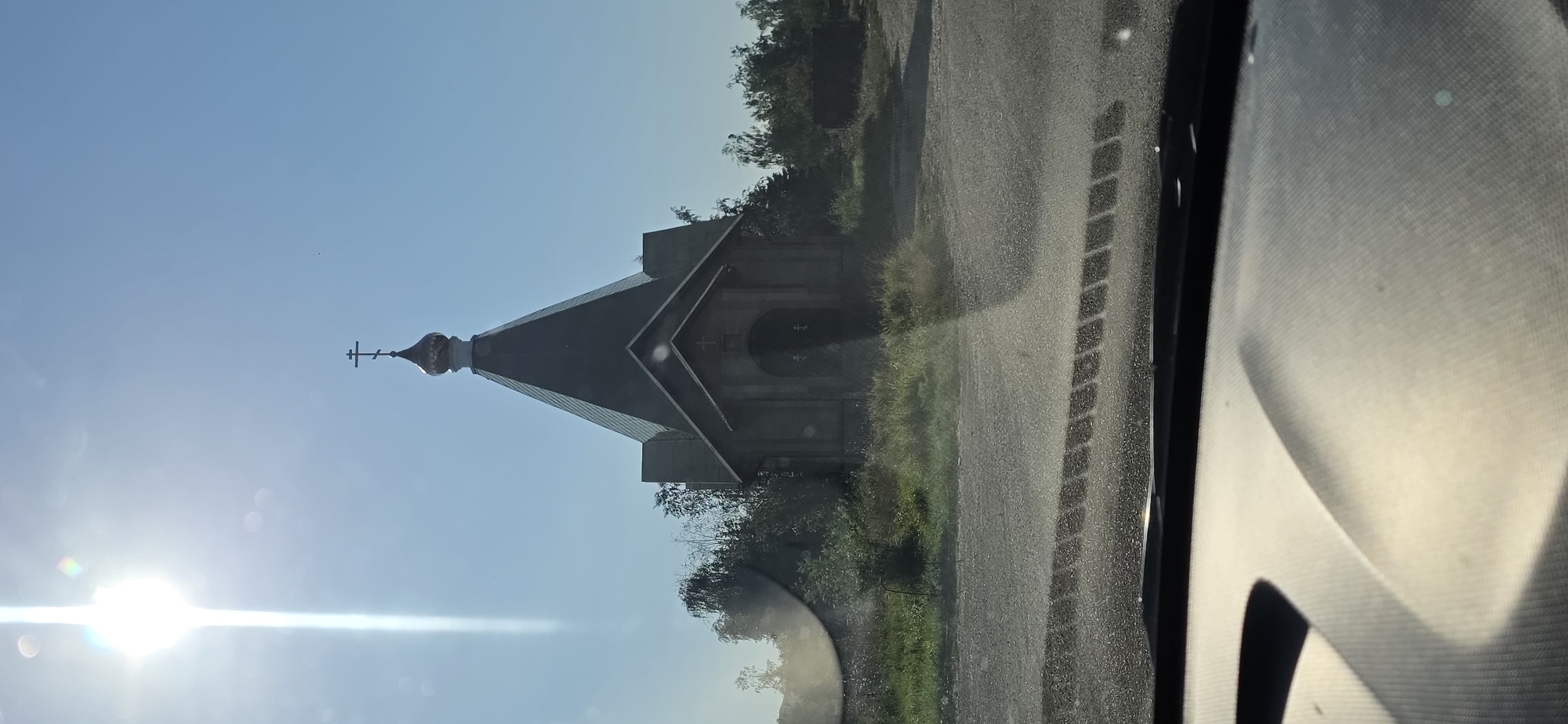 The image shows a small, nondescript chapel with a dark, pyramidal roof topped with a small, golden dome and a cross. The chapel appears to be made of stone or concrete and has an arched entryway. It is situated in a rural or semi-rural setting with trees and overgrown vegetation surrounding it. The ground in front of the chapel is a rough, gravelly surface, possibly a road or parking area. The sun is bright and high in the sky, casting shadows and creating glare on the windshield of a vehicle from which the photo appears to have been taken. The sky is clear and blue. The overall impression is one of solitude and a simple, possibly ancient structure. There is no clear indication of the specific location within the city of Leninsky, Russia.