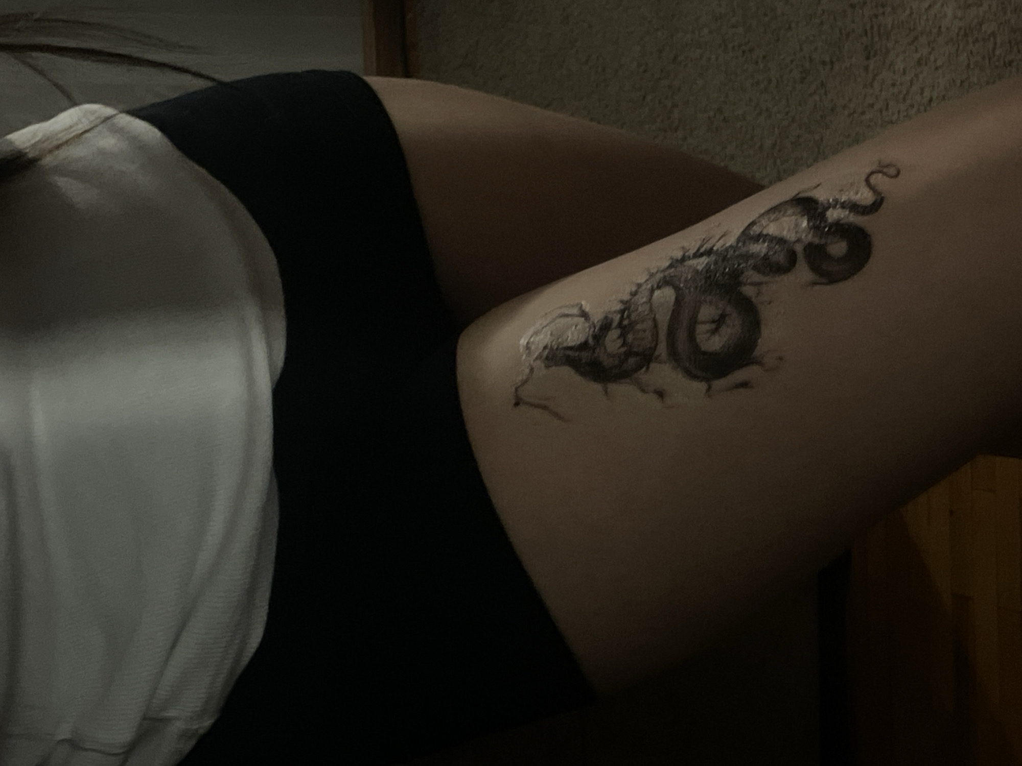 A person's thigh is visible, adorned with a dragon tattoo. The person is wearing black shorts and a white shirt. The background is blurry and dark. The location cannot be determined from the image.