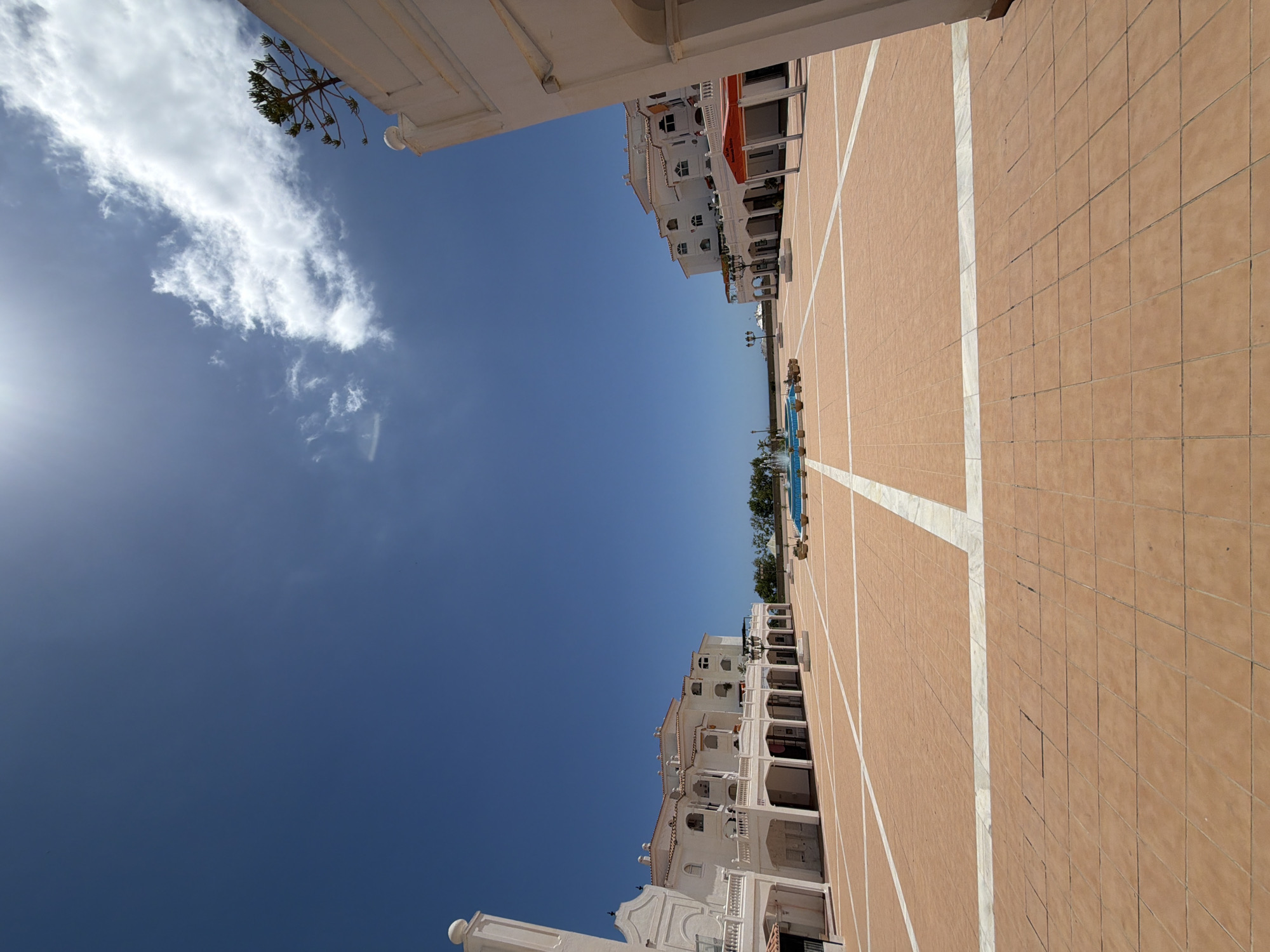The image depicts a view of a public square in Arroyo de la Miel-Benalmádena Costa, Spain. The square is paved with reddish-brown tiles marked with white lines. Flanking the square are white buildings with balconies and arched supports. In the distance, there is a glimpse of a swimming pool with trees and a distant view of the sea. The sky is blue with some scattered white clouds. The sun is visible in the lower left.