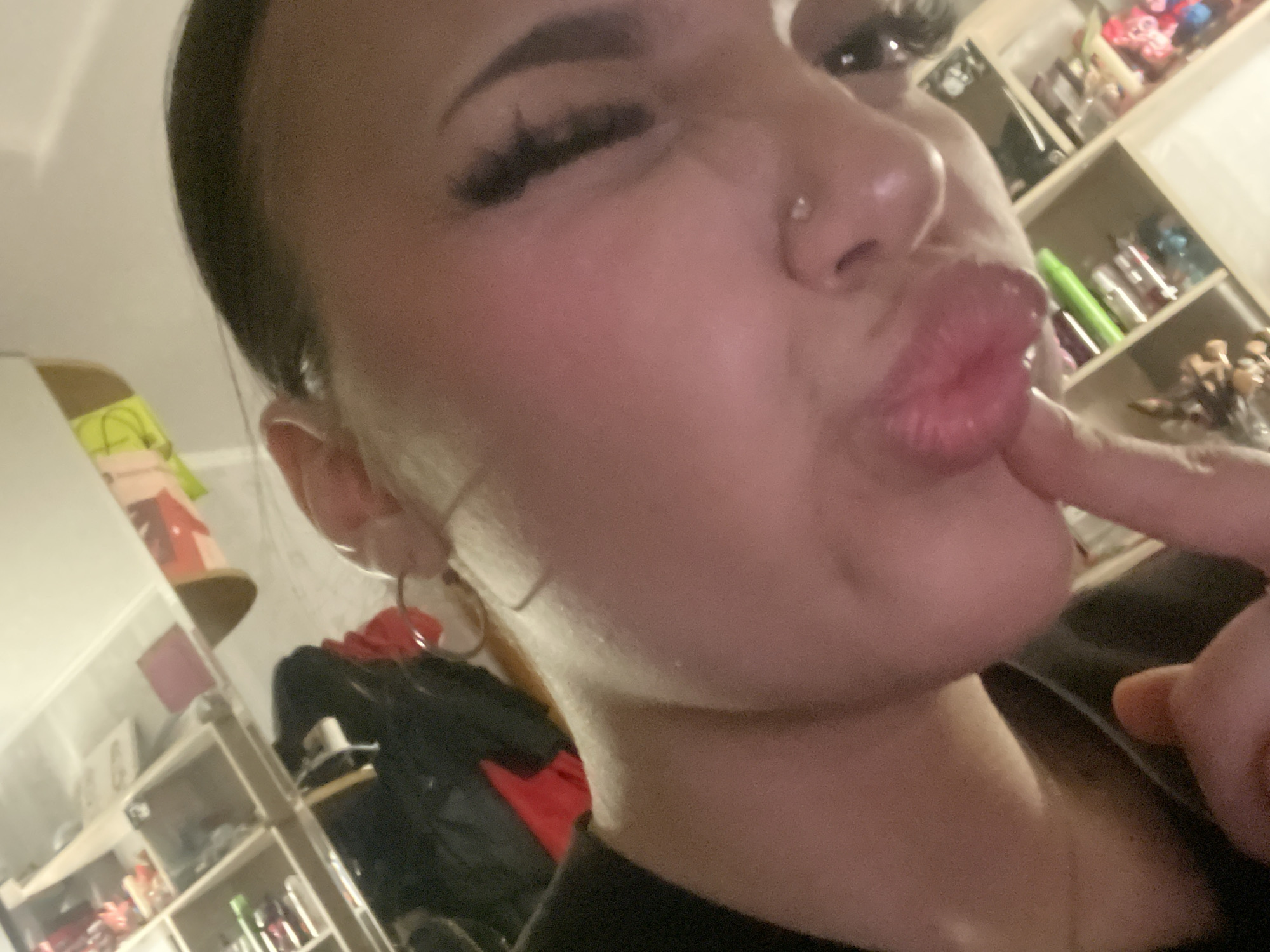 A close-up, angled shot captures a person with fair skin, dark hair pulled back, and prominent eyelashes, posing with pursed lips. A small stud adorns their nose. Their right eye is partially closed in a wink. A finger is placed against their lips. The background features a cluttered shelving unit holding an assortment of items including cosmetics, toiletries, and decorative objects. A garment, predominantly dark with a red element, is draped over something below the shelving. The environment seems domestic. There is no information available to suggest that this image was taken in Murino, Russia.