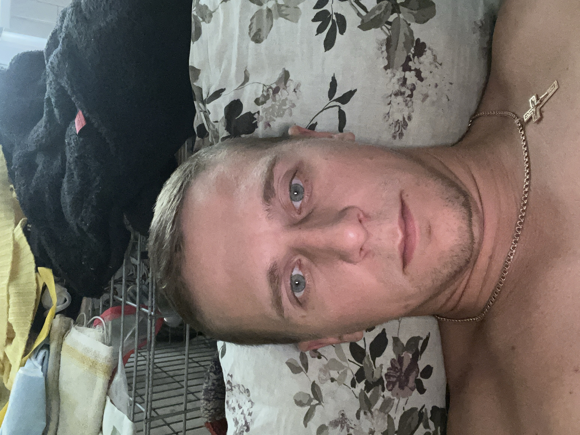 The image is a close-up selfie of a man lying down, with his head resting on a floral patterned pillow. He has short, blond hair and light blue eyes. He is wearing a gold chain with a cross pendant around his neck. Behind him, a metal shopping cart is visible, filled with various items including laundry and blankets. The scene appears to be indoors, possibly in a domestic setting. The location is identified as Minsk, Belarus.