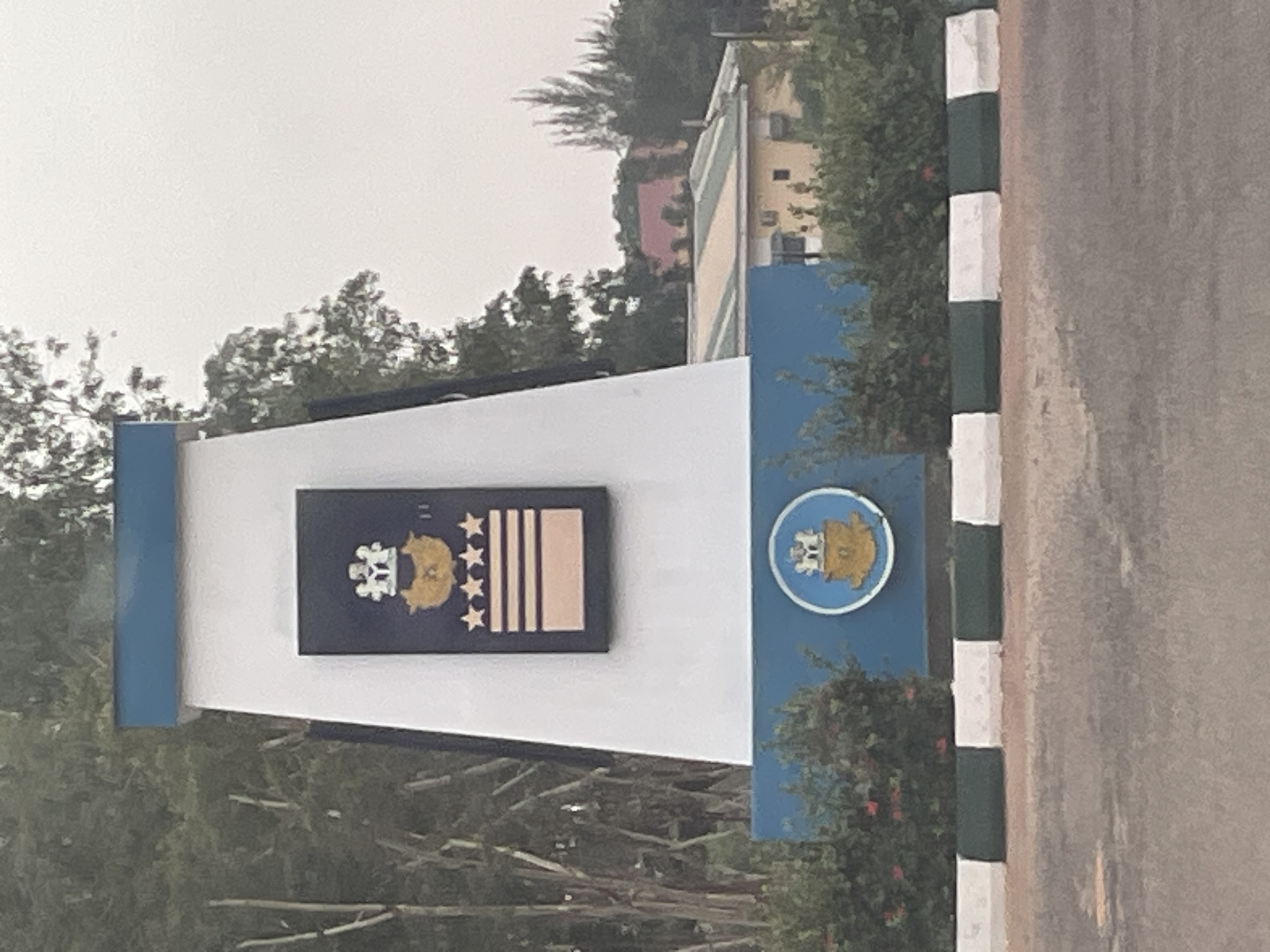 The image shows a sign at the Nigerian Defence Academy in Afaka, Nigeria. The sign features the Academy's emblem and rank insignia. The environment includes trees, shrubs, a building in the background, and a striped curb. The image appears to be taken during the day under overcast conditions. There are no people visible in the frame.