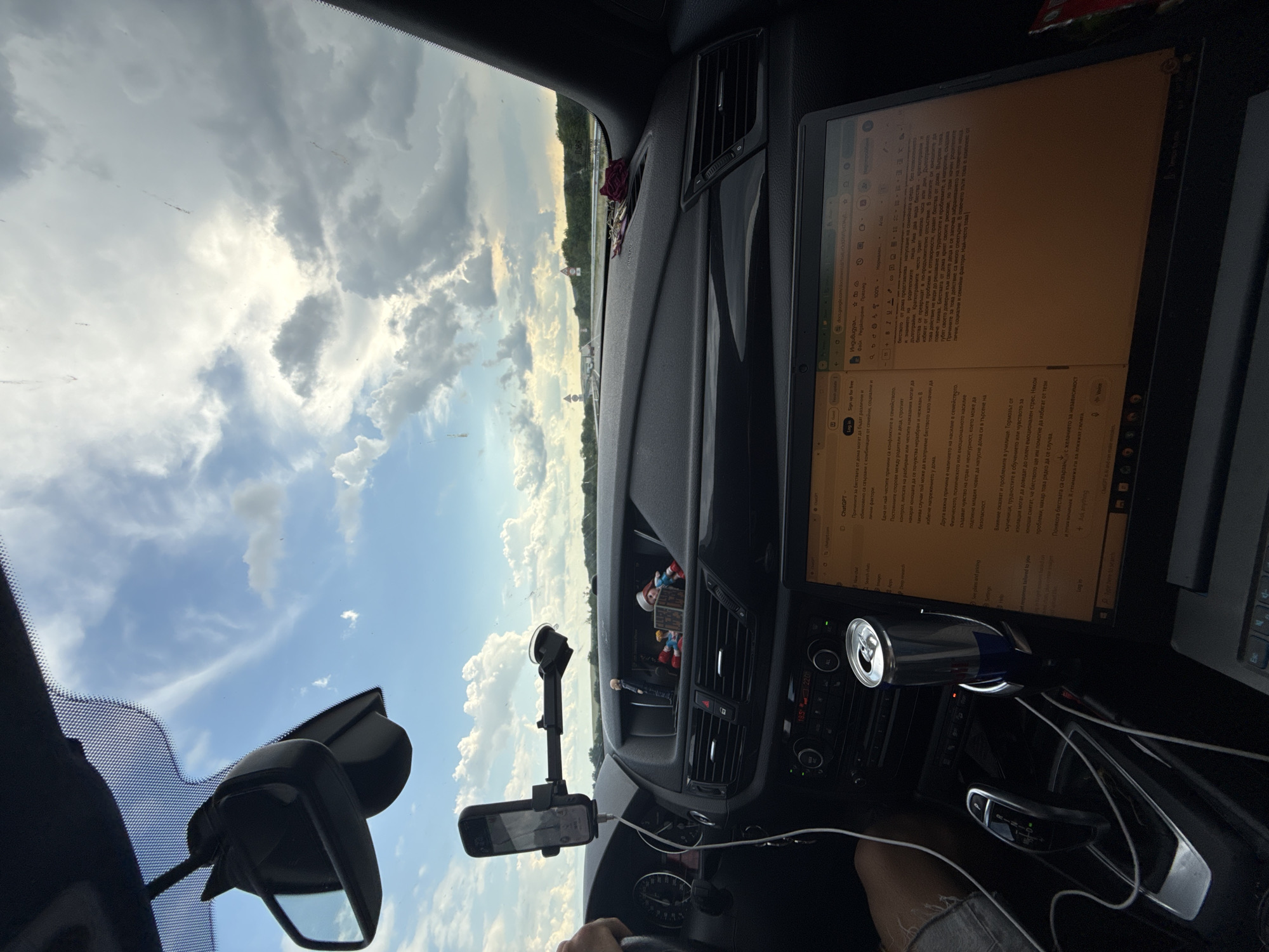 The image shows the interior of a car. A laptop is open on the dashboard, displaying text. A smartphone is mounted on the windshield. There are several small figurines on the dashboard. A can of Red Bull is in a cup holder. The sky is visible through the windshield, with clouds and a bright sun. The car appears to be on a road, possibly a highway, with trees and fields visible in the distance. The location is identified as Arnautito, Bulgaria.
