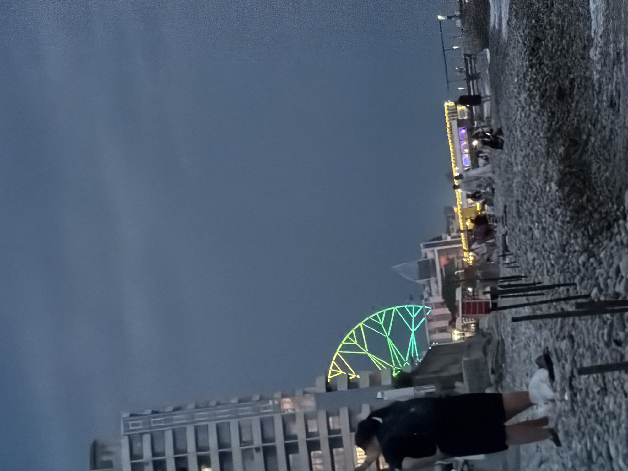 A wide, low-angle shot captures a beach scene at dusk in Sochi, Russia, near a large building and a brightly lit Ferris wheel. In the foreground, a woman in a black dress and white socks stands on a pebbled beach, facing away from the camera, with her hands on her hips. A white object, possibly a bag or garment, lies on the ground near her feet, along with a dark hat. In the middle ground, a cluster of people are gathered near illuminated structures and a Ferris wheel, which is illuminated with green and yellow lights. The background features a tall, multi-story building on the left, its windows reflecting the twilight sky. A long, low building with strings of yellow lights is visible further back, and a body of water can be seen on the right. The sky is a deep, overcast blue.