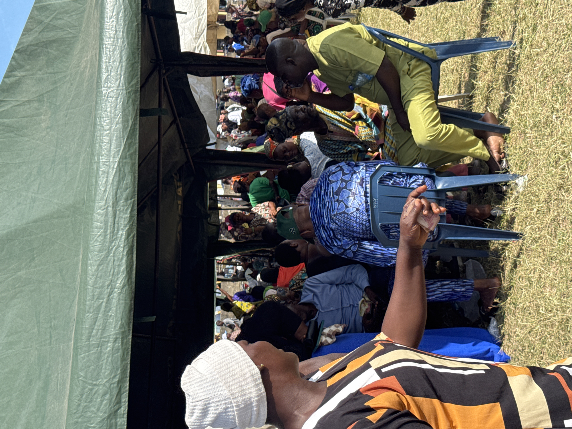 The image shows a large group of people gathered under a green tent in Badagry, Nigeria. They appear to be seated and listening, possibly attending an event or meeting. The people are wearing a variety of colorful clothing. A person in the foreground is wearing a patterned shirt and a white cap. Many of the people are sitting on blue plastic chairs. The event takes place on a grassy area.