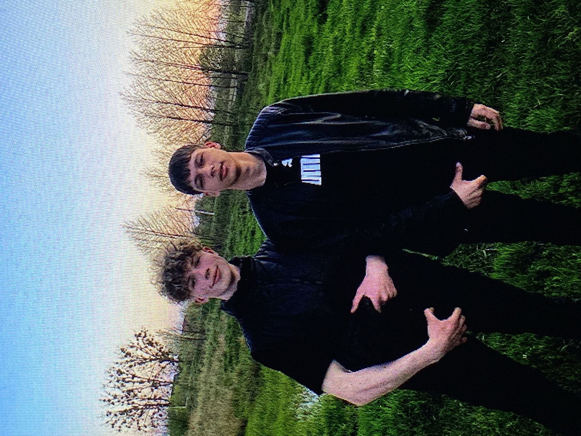 Two young males are standing in a grassy field with trees in the background. The male on the left has curly light brown hair and is wearing a black puffer vest over a black shirt. He is holding his right hand to his stomach. The male on the right has short dark hair and is wearing a black leather jacket over a black t-shirt with a white logo. His left hand is in his jacket pocket. The sky is a pale blue with hints of orange and yellow, suggesting it is either sunrise or sunset. The location is the city of Poznan, Poland.