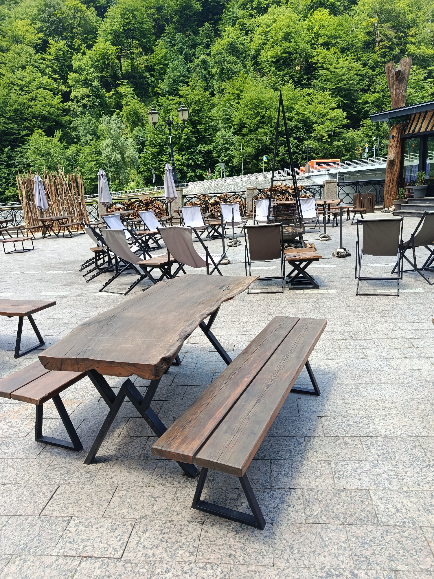 The image depicts an outdoor seating area with wooden tables and benches in Estosadok, Russia. In the foreground, a large, rustic wooden picnic table with an accompanying bench is prominent. Behind this, other tables and chairs are arranged on a paved surface. The background features a dense, green forest on a mountainside, with a road and a bridge visible in the mid-ground. Several street lamps are also present. The scene appears to be a public area, likely a restaurant or cafe's outdoor section, set against a natural backdrop. There are no people visible in the image.
