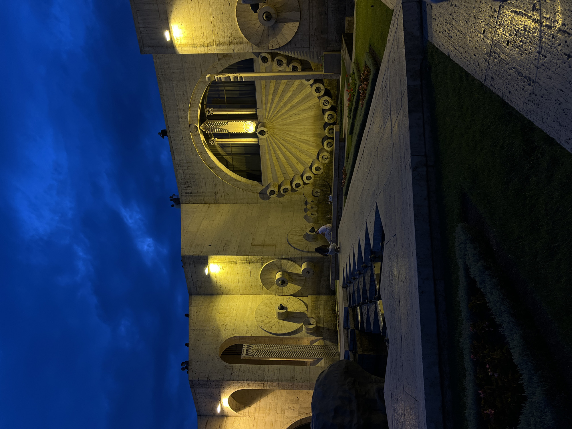 The image depicts a nighttime view of the Cascade in Yerevan, Armenia. The structure is illuminated by warm yellow lights, casting long shadows and highlighting the textured stone architecture. In the foreground, a pathway made of large, light-colored stone slabs leads towards the cascade. To the left, a dark, sculpted object, possibly a fountain head, is visible. Several people are present; some are seated on the steps of the cascade, while others are visible on the upper levels of the structure, silhouetted against the deep blue, cloudy sky. The cascade itself features a large, circular element with radiating lines and decorative elements, as well as several large, wheel-like sculptures on the surrounding walls. Flower beds with low-lying plants and occasional red flowers are visible along the pathway, contrasting with the stone and grass. The overall atmosphere is one of quiet evening illumination of a significant architectural landmark.