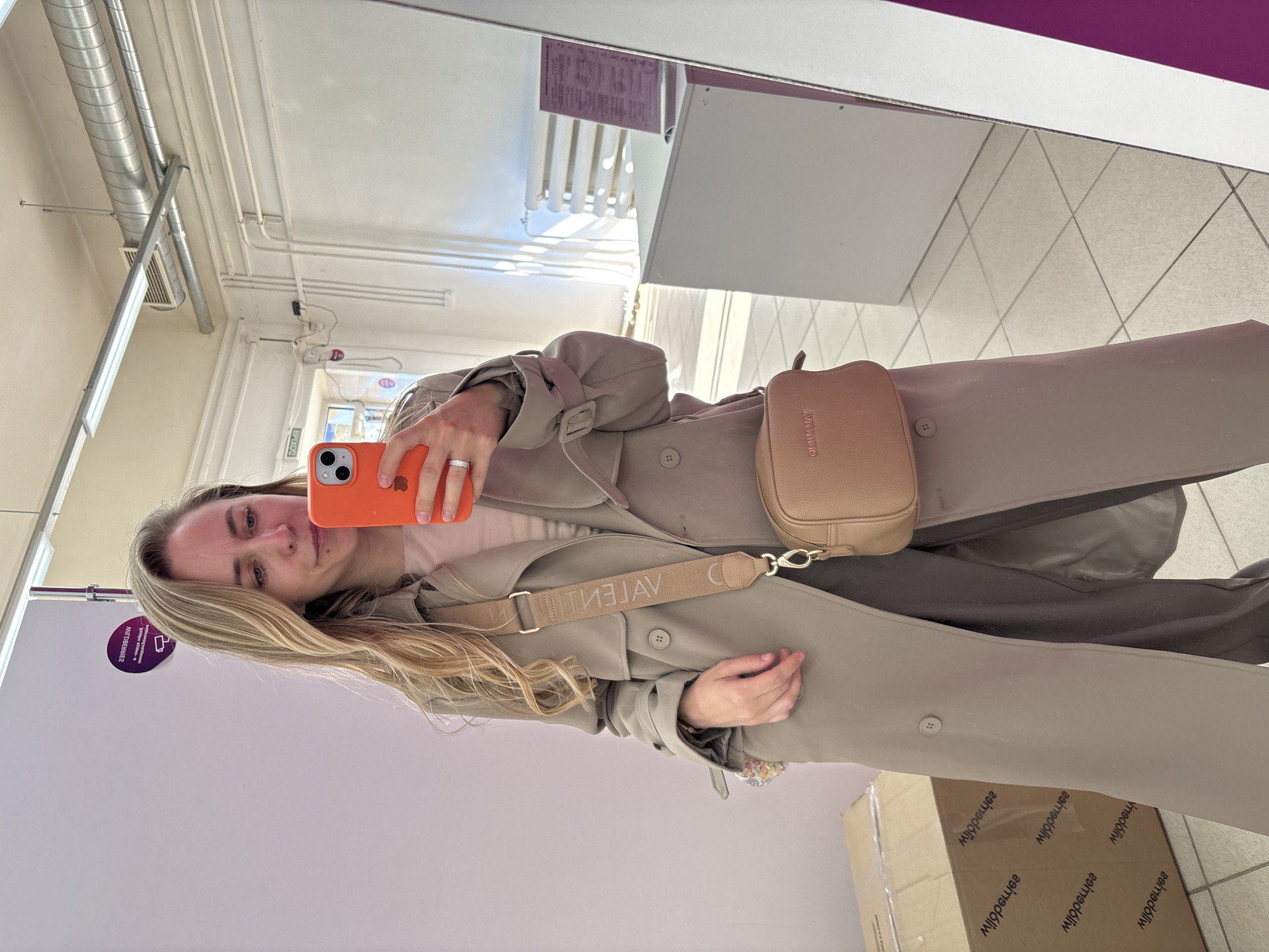 A blonde woman is taking a selfie in front of a mirror. She is wearing a beige trench coat and a beige shoulder bag with the brand name "VALENTINI" printed on the strap. She is holding an orange iPhone.
Based on textual data on an environmental sticker, the image was taken in the city of Chaikovsky, Russia.
The reflection in the mirror shows a retail or commercial setting, with lighting fixtures on the ceiling and a tiled floor. There is also a tall white cabinet visible in the reflection.