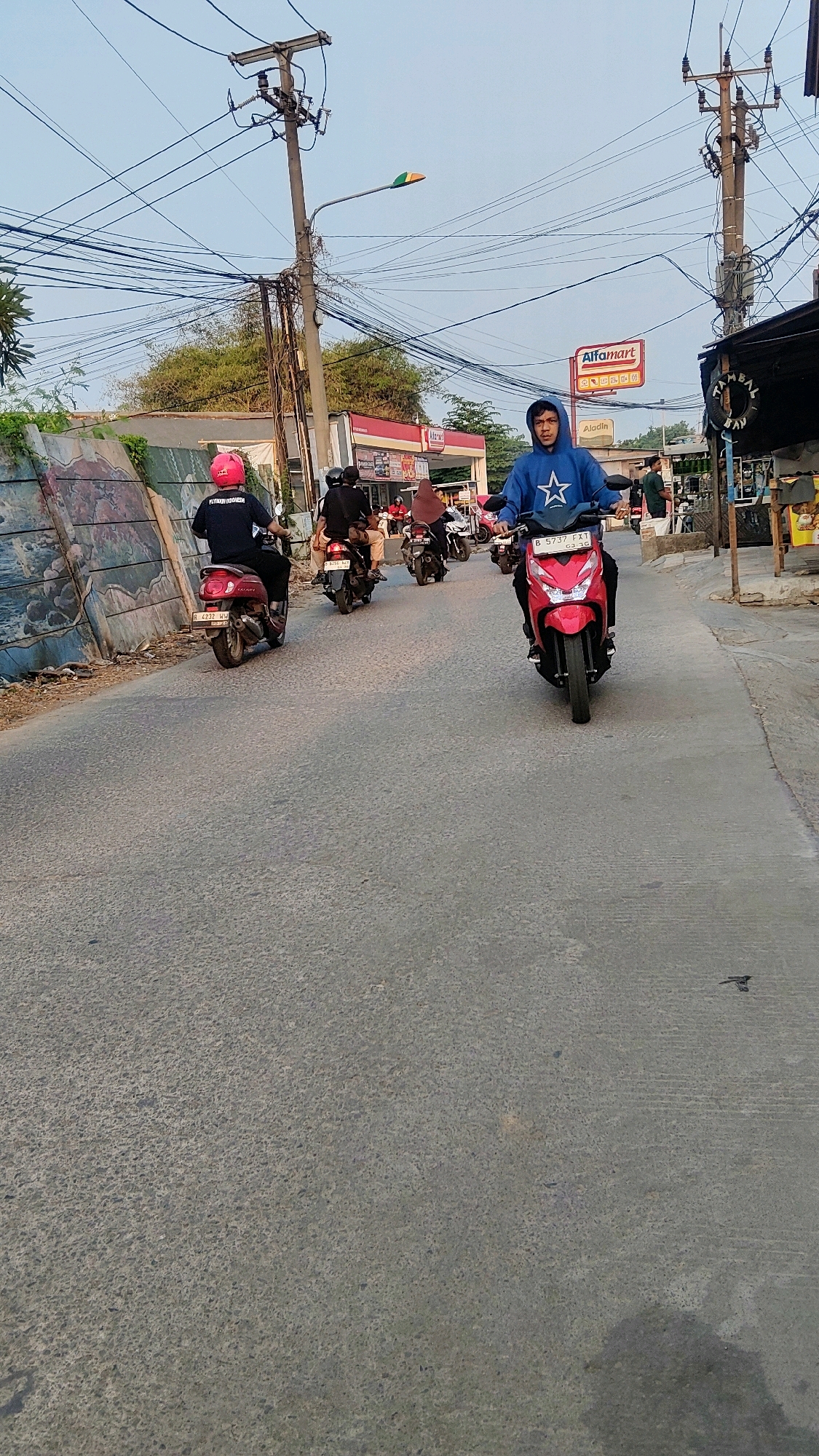 A street scene in Mekarmukti, Indonesia, depicts several individuals riding motorcycles on a paved road. In the foreground, a person wearing a blue hooded sweatshirt and a white star emblem is riding a red motorcycle. Behind them, a person in a pink helmet on a red motorcycle is visible. Further back, other motorcyclists are present, with one wearing a black hijab. The street is lined with utility poles carrying numerous electrical wires, a street light with multi-colored panels, and commercial establishments including an Alfamart. The ground is asphalt, showing wear and tear. Vegetation is present on the left side of the image, alongside a graffiti-covered wall. The sky is clear and light blue, suggesting it is daytime.