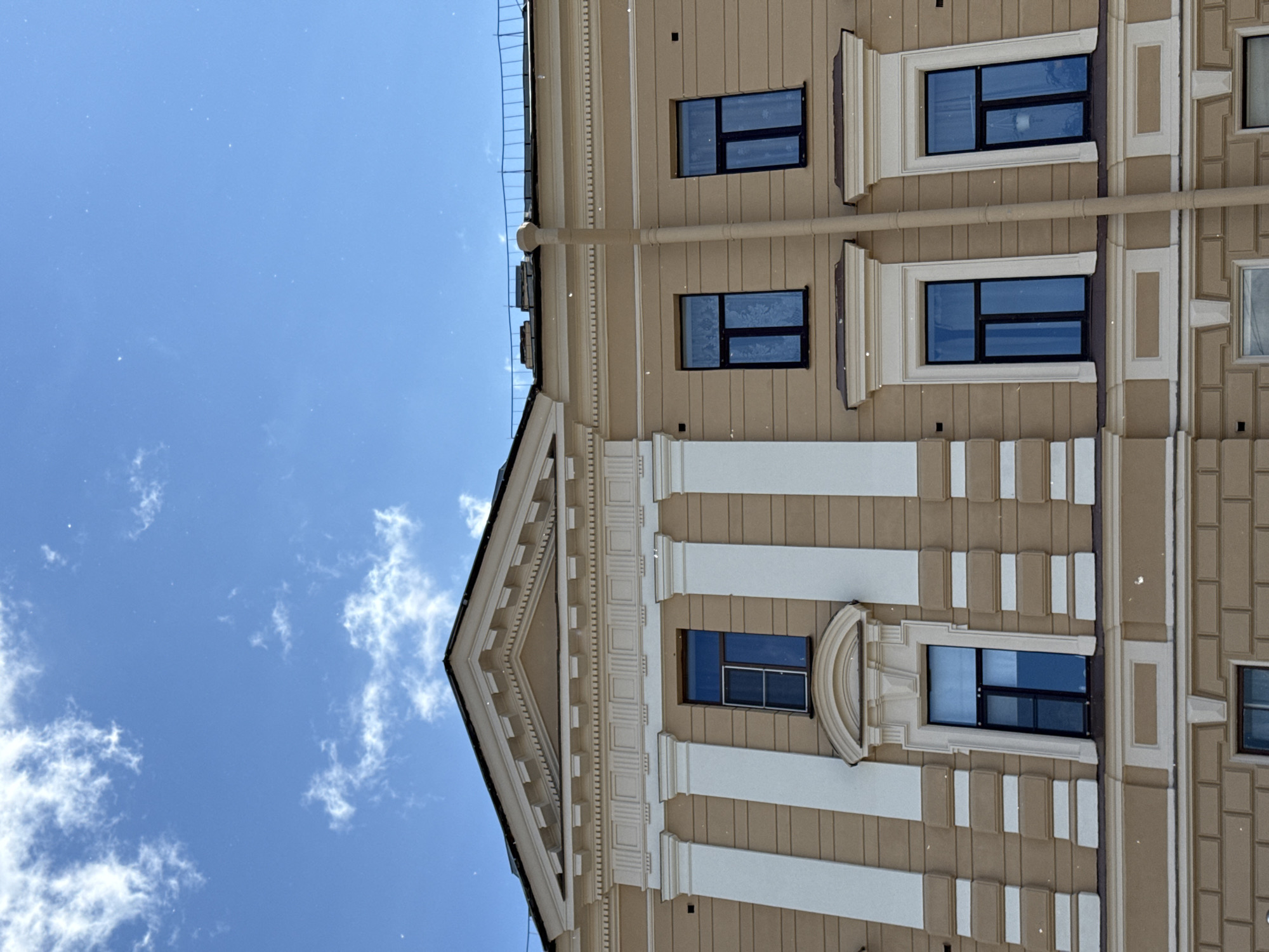 This is a photograph of the facade of a building in Saint Petersburg, Russia, under a bright blue sky with scattered white clouds. The building is a light beige color with white decorative columns and horizontal banding. There are several rectangular windows with dark frames and blue-tinted glass. Some of the windows have decorative white surrounds. A prominent downspout runs vertically along the center right of the building. The architecture suggests a historical or classical style, common in Saint Petersburg. The image is taken from a low angle, looking upwards towards the sky and the upper floors of the building.