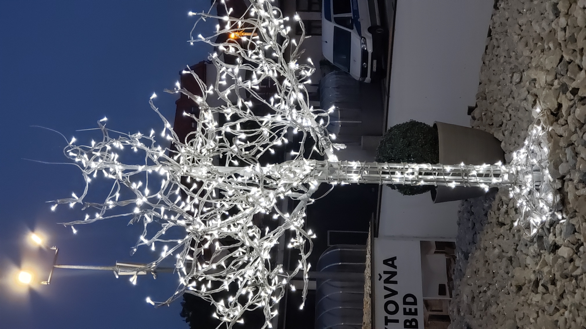 This is a vertical, medium-shot photograph taken at dusk in Prešov, Slovakia, depicting a decorative tree illuminated by numerous white LED lights. The tree's structure, made of white material, is visible through the lights, which are wound around its branches and trunk. The ground around the tree is covered in light-colored gravel, also illuminated by some of the lights. To the left of the tree, a portion of a white sign with black lettering is visible, reading "VTOVŇA BED". Behind the tree, a large, dark gray pot contains a spherical, green topiary plant. Further back, a white van with a blue stripe is parked. The background shows a dark blue sky with a streetlamp visible in the upper left corner, and hints of buildings with red roofs. The overall impression is of an outdoor decorative display at night, likely in a commercial or public area.
