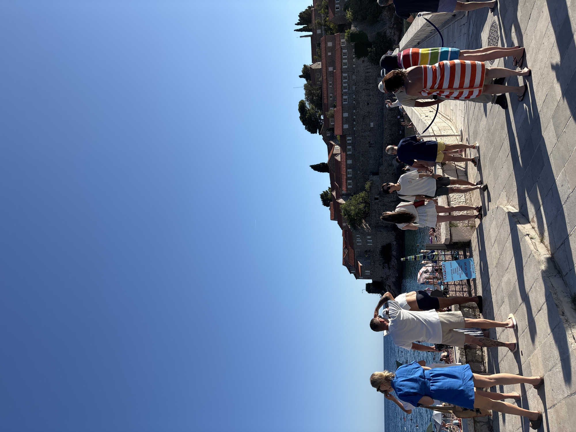 In Sveti Stefan, Montenegro, a group of people are on a pathway leading towards the historic island. The pathway is paved with light-colored stones, and a low stone wall with a rope railing lines the right side. The island, visible in the background, is densely packed with stone buildings topped with red tile roofs. Lush green trees are scattered among the buildings.
In the foreground, several individuals are walking and standing. A woman in a bright blue dress is on the left, facing away from the camera. To her right, a man in a white t-shirt and tan shorts is looking out towards the sea. Behind him, other people are also visible on the pathway.
Beyond the pathway and to the left, a beach with numerous umbrellas and sunbathers is visible along the shoreline. The sea is a deep blue, meeting the clear, bright blue sky. A sign with "TAXI BOAT" written on it is propped up near the beach entrance. The lighting suggests it is a sunny day, with distinct shadows cast by the people and the wall.