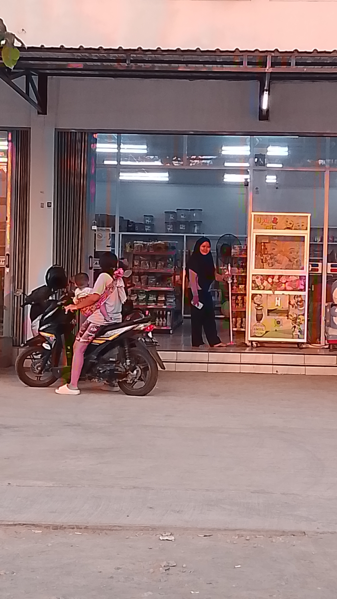 In Karanglangu, Indonesia, a person is seated on a motorcycle, holding a baby. The person wears a white t-shirt and patterned shorts, with pink leggings visible. The baby is secured in a patterned sling. The motorcycle is black and has a yellow and black decal on its side. In the background, a woman wearing a black hijab and black attire is standing near a display rack filled with various items. Behind her, shelves stocked with products are visible inside a building. The scene takes place on a paved area in front of a building with large glass windows and an overhang.