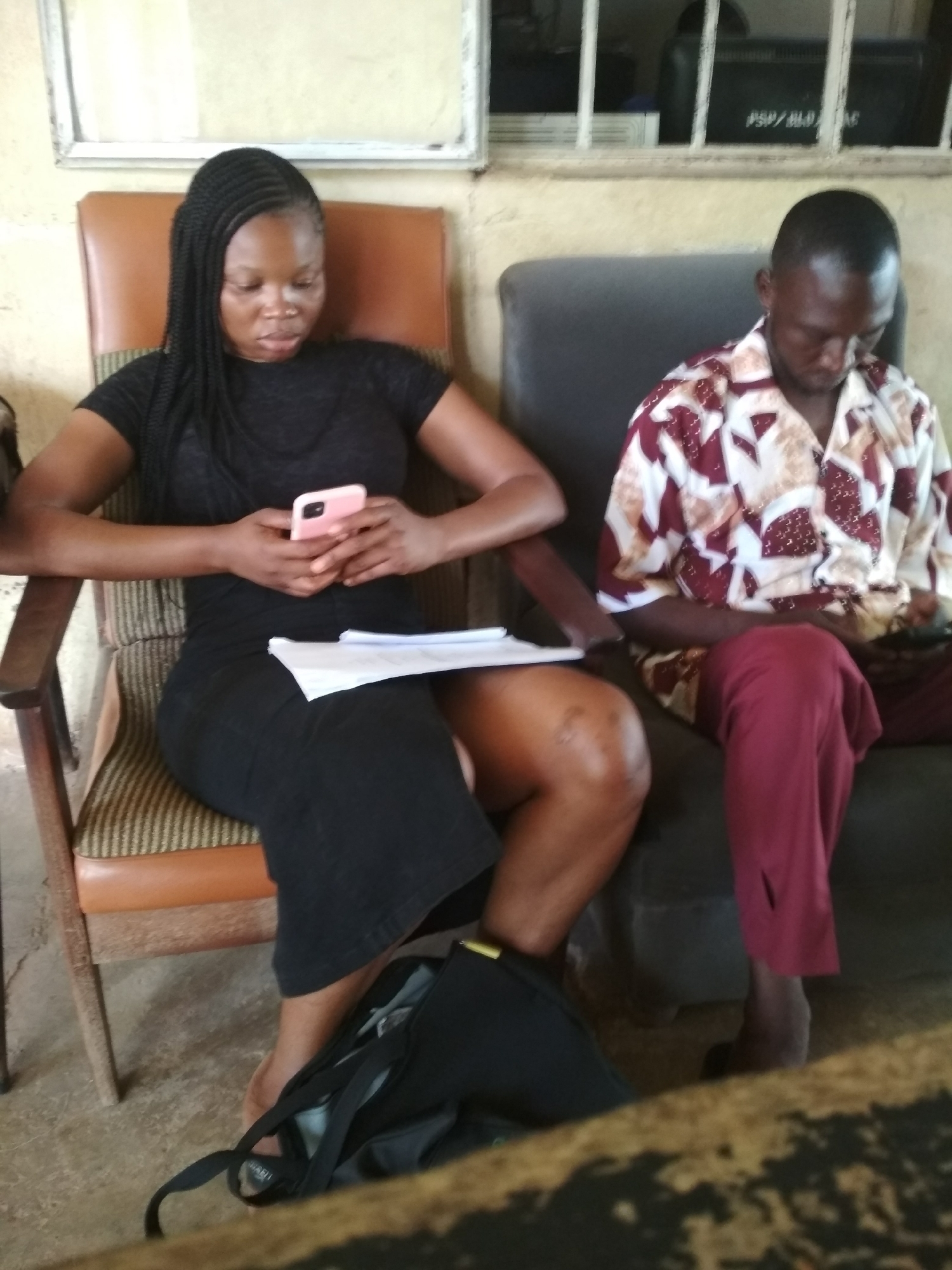 Two individuals are seated in chairs, appearing to be in a waiting area or common room. On the left, a woman with braided hair is wearing a black t-shirt and a black skirt. She is holding a pink mobile phone in her hands and looking down at it. A piece of paper rests on her lap, partially obscured by her clothing. A black bag is on the floor next to her. To her right, a man wearing a maroon and white patterned shirt and maroon pants is seated, looking down at something in his lap. The environment appears to be indoors, with walls in the background and furniture consisting of chairs. A window with bars is visible in the background. There is no specific information in the image to indicate the location as Bisichi, Nigeria.