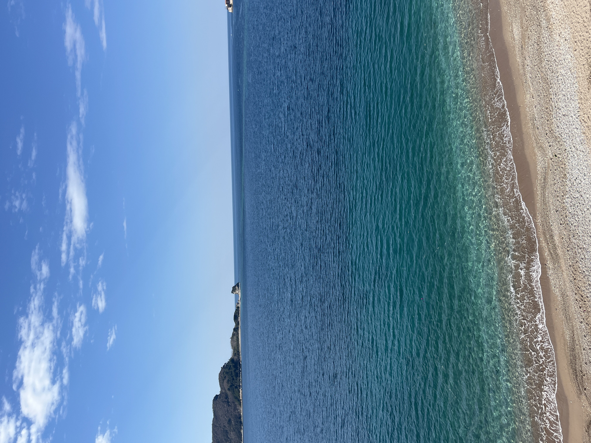 The image shows a beach scene in Budva, Montenegro. The sky is blue with scattered white clouds. The sea is a gradient of blue, from deep navy further out to a turquoise near the shore. A rocky cliff borders one side of the sea. The beach is sandy with small pebbles.