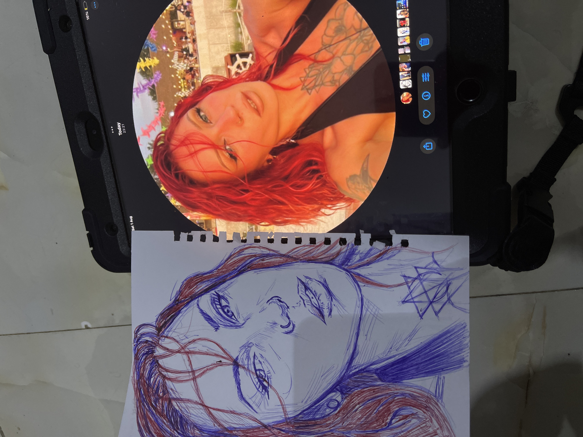 A pen sketch of a woman with red hair is placed next to an iPad displaying a circular photograph of the same woman. The sketch is on a white piece of paper with torn edges, and it is rendered in blue and red ink. The woman in the sketch has her head tilted, and her facial features, including her eyes, nose, and mouth, are detailed. Red lines are used to depict her hair. There is a geometric symbol drawn in blue ink below her neck.
The iPad shows a photograph of the woman with vibrant red hair, wearing a black top. She has a tattoo on her chest. The photograph is set against a background of an outdoor venue with colorful decorations. The time displayed on the iPad is 23:21, and the date is Today, August 1st. A battery indicator shows 16% charge. Below the photograph, there are icons for sharing, liking, information, and deleting. The iPad is placed on a surface that appears to be a tiled floor. The overall scene is captured from a slightly overhead perspective. There is no indication of the city of Abuja, Nigeria, in the image.