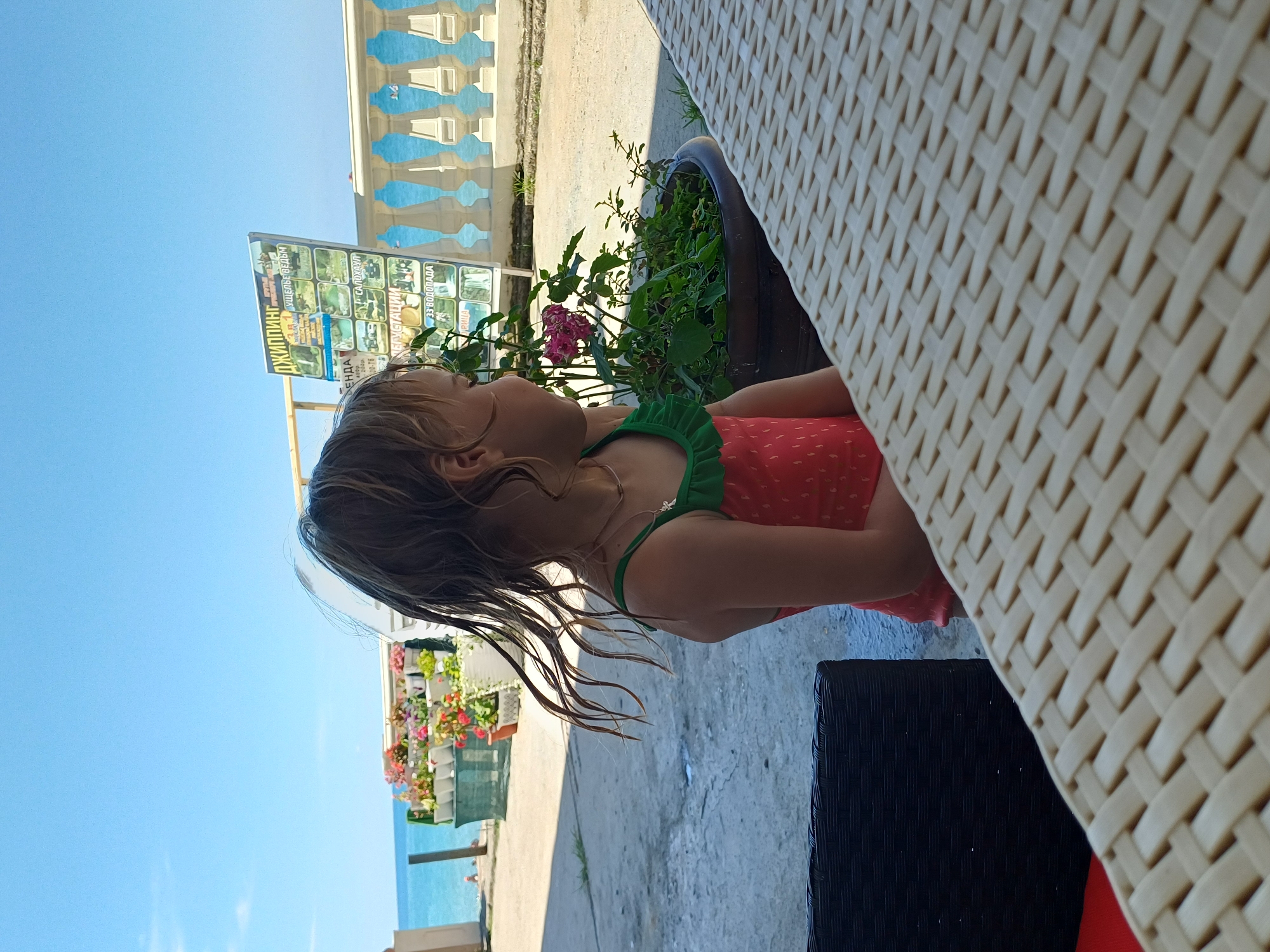 This is a vertical, close-up, eye-level shot of a young girl sitting at a table outdoors. The girl has blonde hair and is wearing a red swimsuit with white polka dots and a green ruffled neckline. She is looking up and to the left, with a slight smile. Her hair appears wet and is falling around her face and shoulders.
The setting is outdoors, with a bright blue sky visible. To the left, there is a white building with decorative blue railings, suggesting a coastal or resort location. Below the building, there are colorful flowers in planters and a glimpse of the sea. A signpost with several pictures and text in Russian is also visible, advertising various activities, including "ДЖИППИНГ" (Jeeping), "КАНЬОНИНГ" (Canyoning), and "33 ВОДОПАДА" (33 Waterfalls).
The table the girl is sitting at has a woven texture, possibly made of rattan or wicker, and part of another table or chair made of black woven material is visible in the foreground. The time of day appears to be daytime, with bright sunlight casting shadows. The overall mood of the image is relaxed and summery, evoking a vacation or leisure setting.