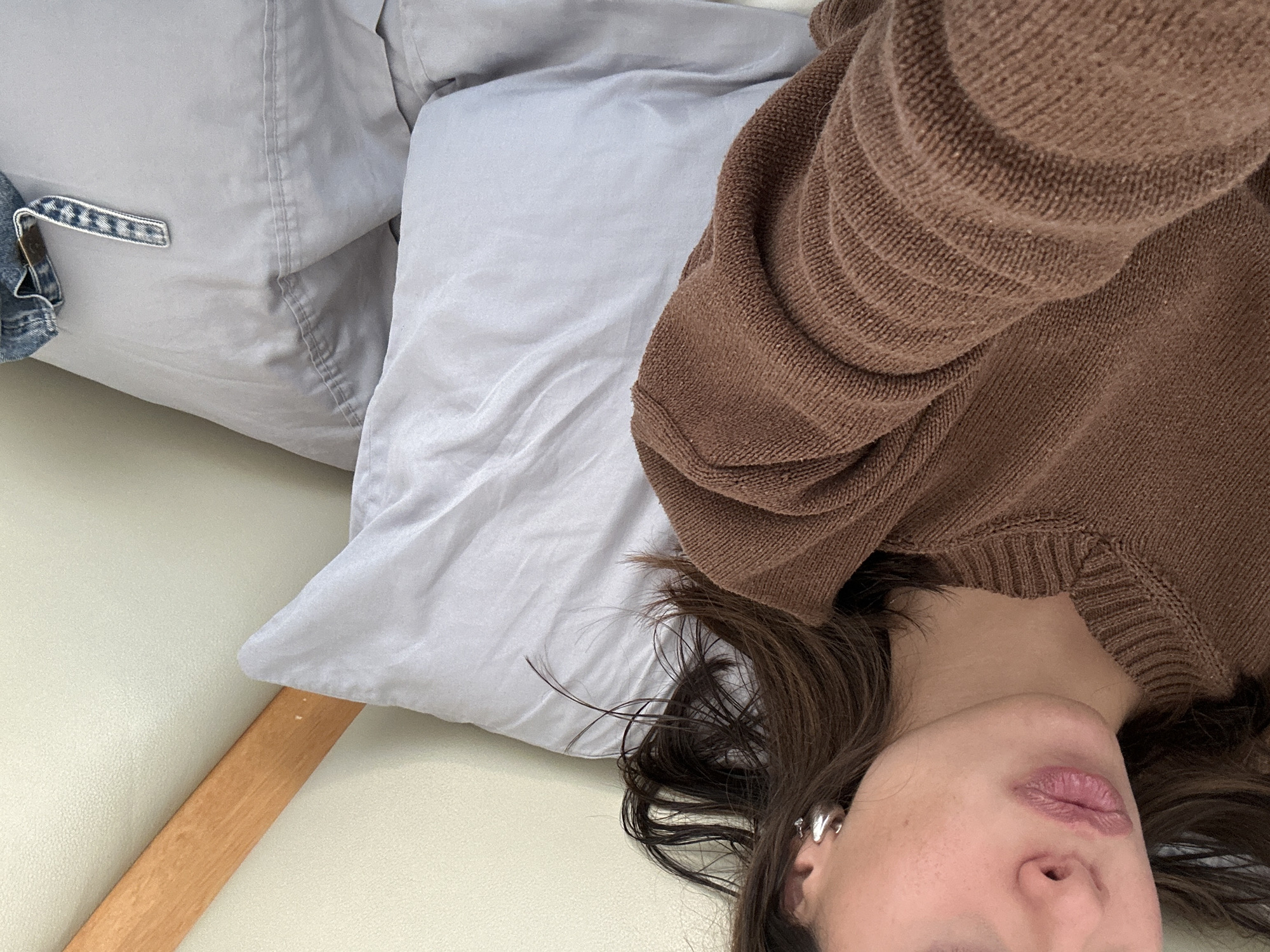 A person wearing a brown sweater is lying on a light gray pillow. The pillow is on a light-colored surface next to a wooden chair leg. Part of a denim jacket is visible on the left side of the image. The location is Unknown, Unknown.