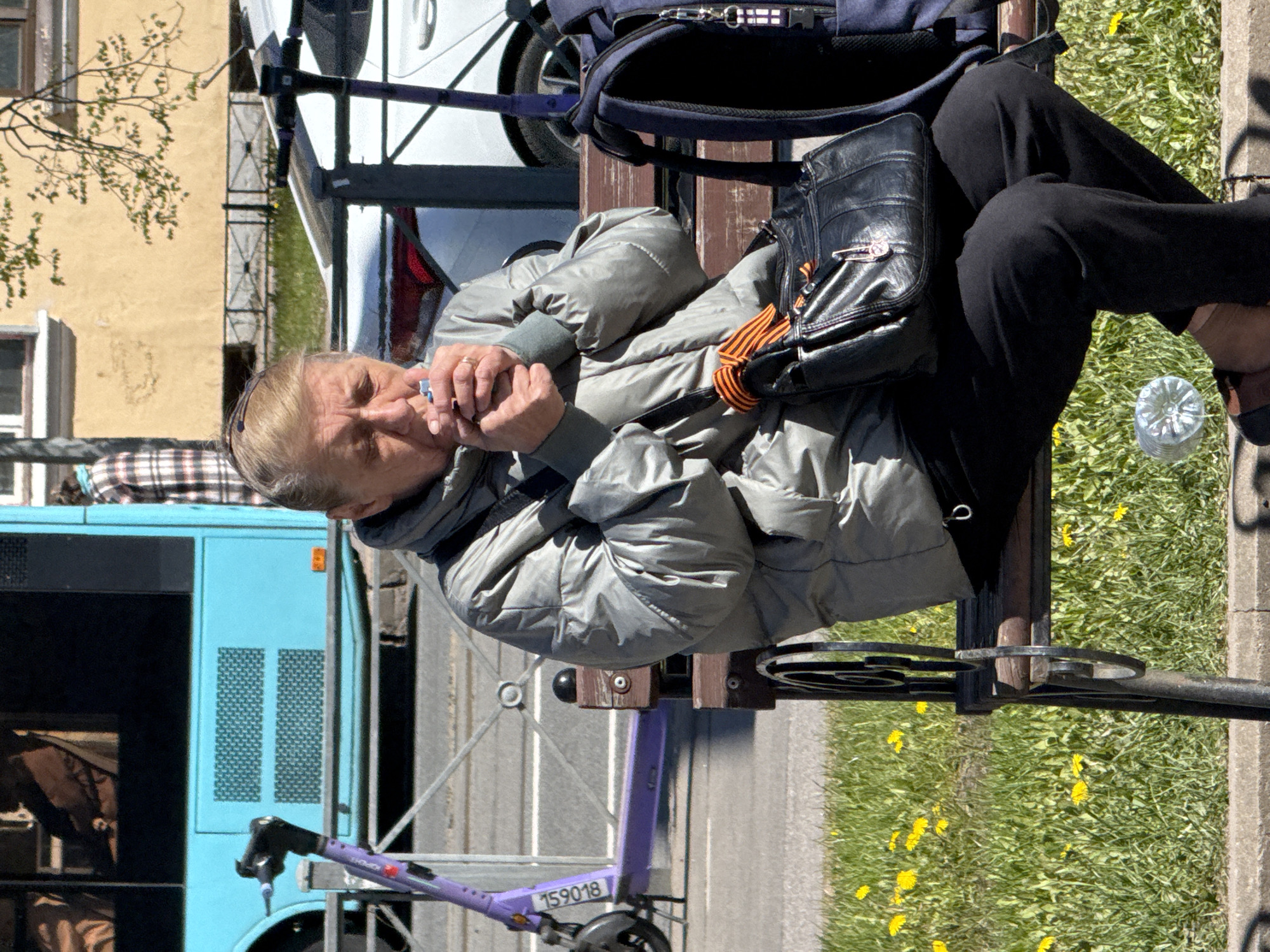 An elderly person sits on a park bench in Saint Petersburg, Russia, smoking. The individual wears a grey puffer jacket and black pants and carries a black leather bag with an orange ribbon. A blue bus and a purple bicycle share can be seen in the background. The bench is positioned near a sidewalk and grassy area with dandelions. A water bottle sits nearby.