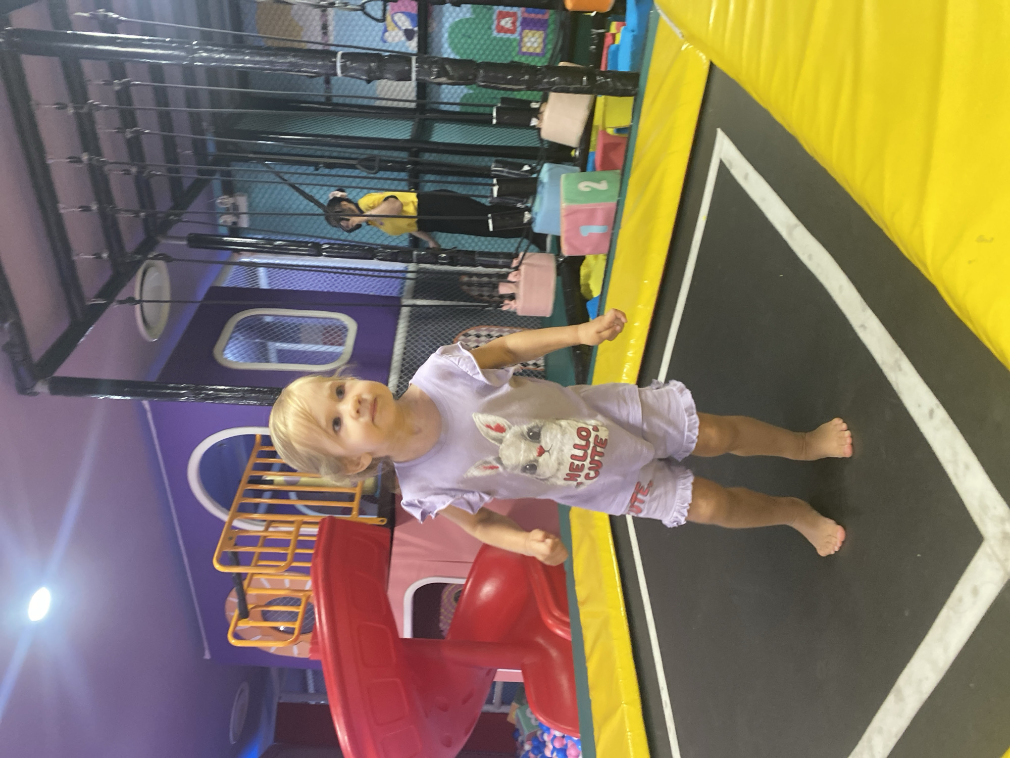 A young girl with blonde hair is standing on a black trampoline with yellow padding in an indoor play area. She is wearing a light purple t-shirt with a rabbit graphic and the words "HELLO CUTE" and matching shorts. The background features a red slide, purple walls, and various play structures. In the background, a woman in a yellow shirt and black pants is visible. The location is identified as Phu Thuy, Vietnam.