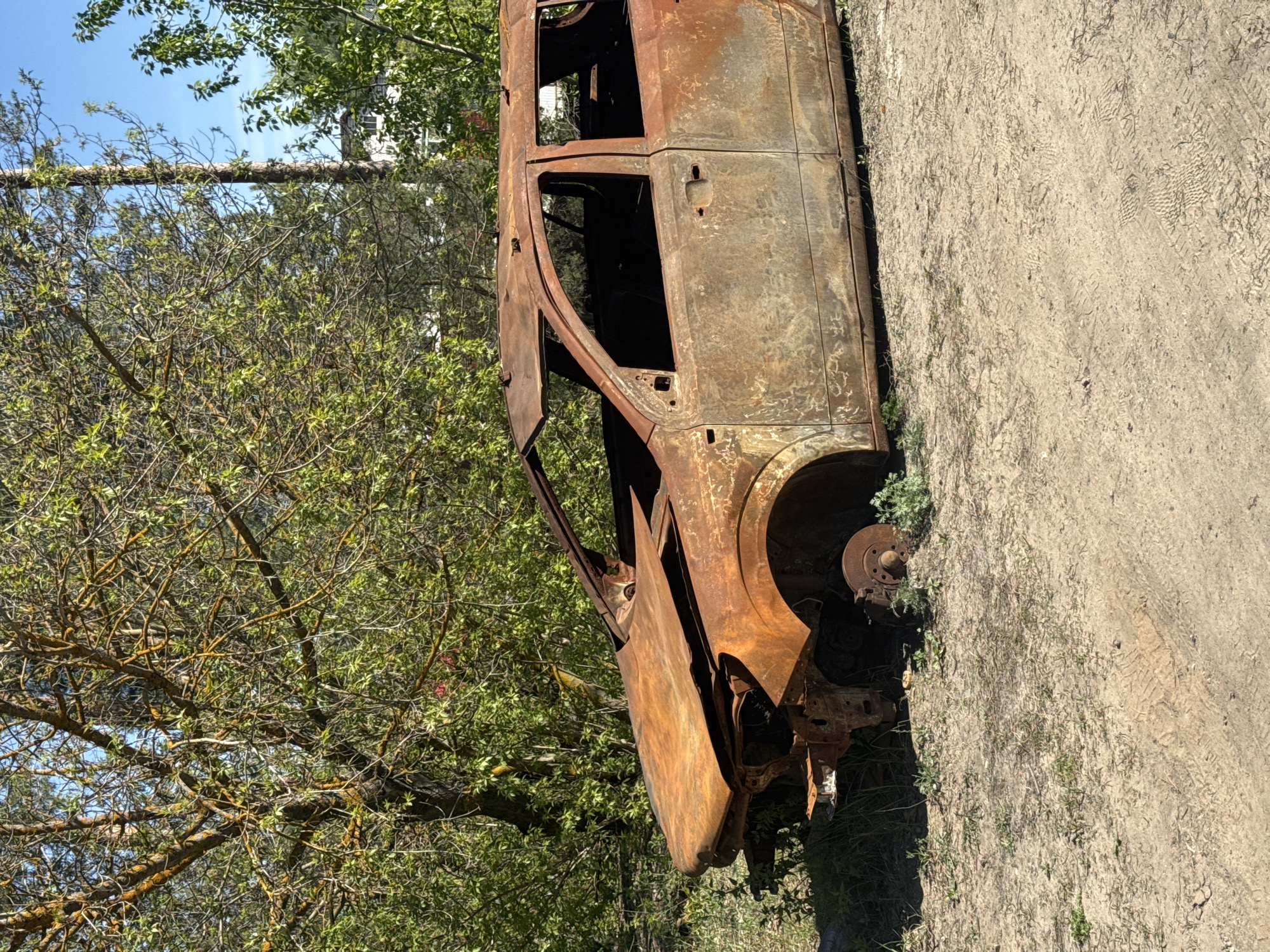 A rusted, burned-out car frame is located on a dirt road next to green trees. The car is heavily damaged with missing doors, windows, and tires. The road shows tire tracks. The specific location in Kazan, Russia, is not identifiable from this image. There are no people visible.