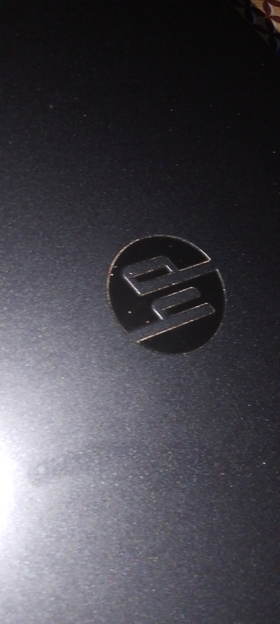 The image shows a close-up view of an HP logo on a dark, textured surface, likely a laptop lid. The logo is circular with the stylized "hp" letters inside, rendered in a metallic or reflective material that stands out against the matte background. The surface appears to be a dark gray or black and shows some subtle texture and a few specks of dust or debris. The lighting creates some highlights on the logo and the textured surface, particularly on the left side of the image where a bright, out-of-focus light source is visible. The image is tightly cropped around the logo, providing no context about the surrounding environment or location.