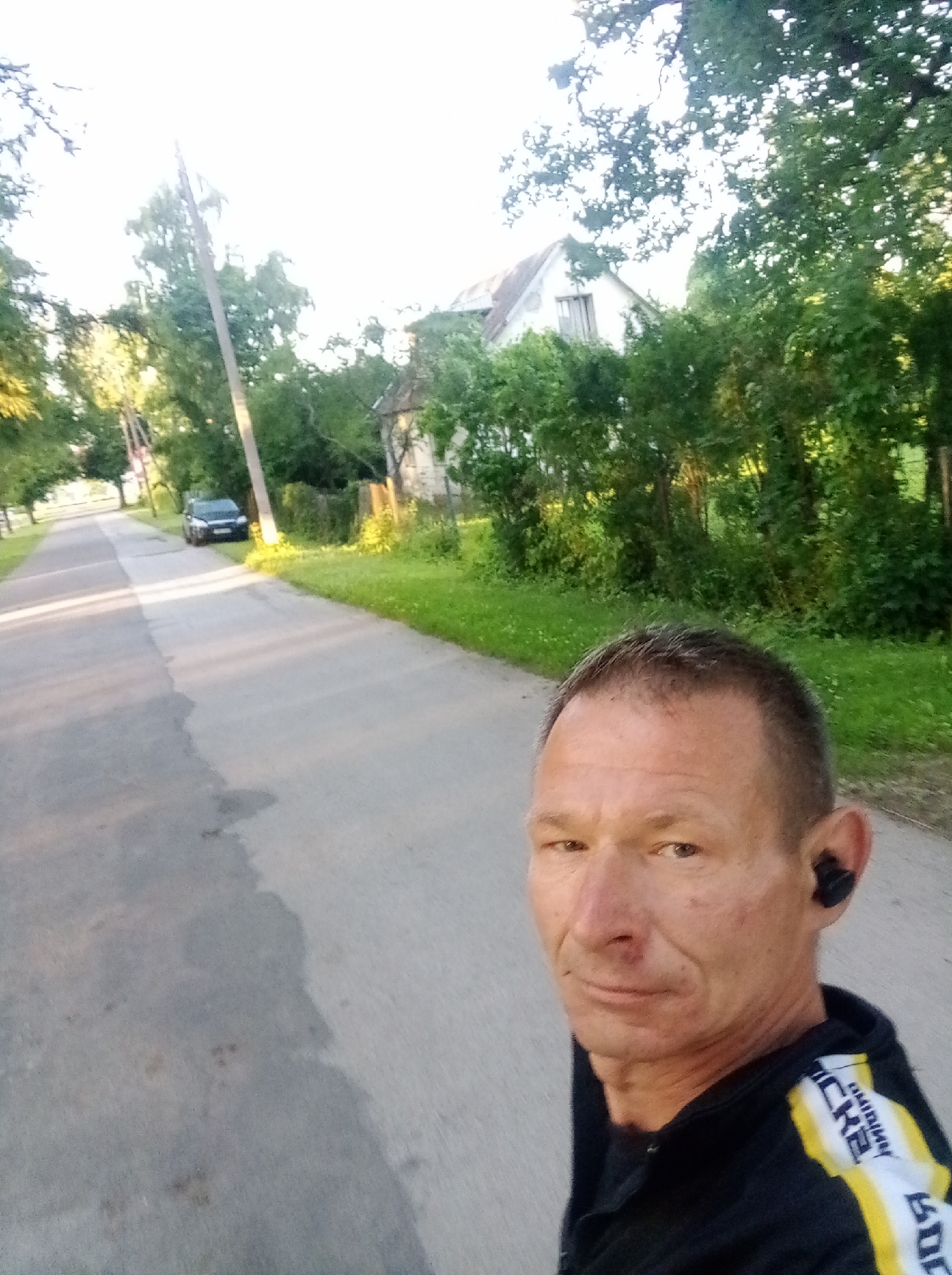 The image depicts a selfie taken by a man with short grey hair and fair skin, wearing black earbuds. He is wearing a black jacket with yellow and white stripes on the sleeves. He is standing on a paved road in a rural or suburban area. In the background, there is a line of trees and a white house with a grey roof. A dark colored car is parked on the side of the road to the left. The road appears to be in Tapa, Estonia. The lighting suggests it is late afternoon or early evening.