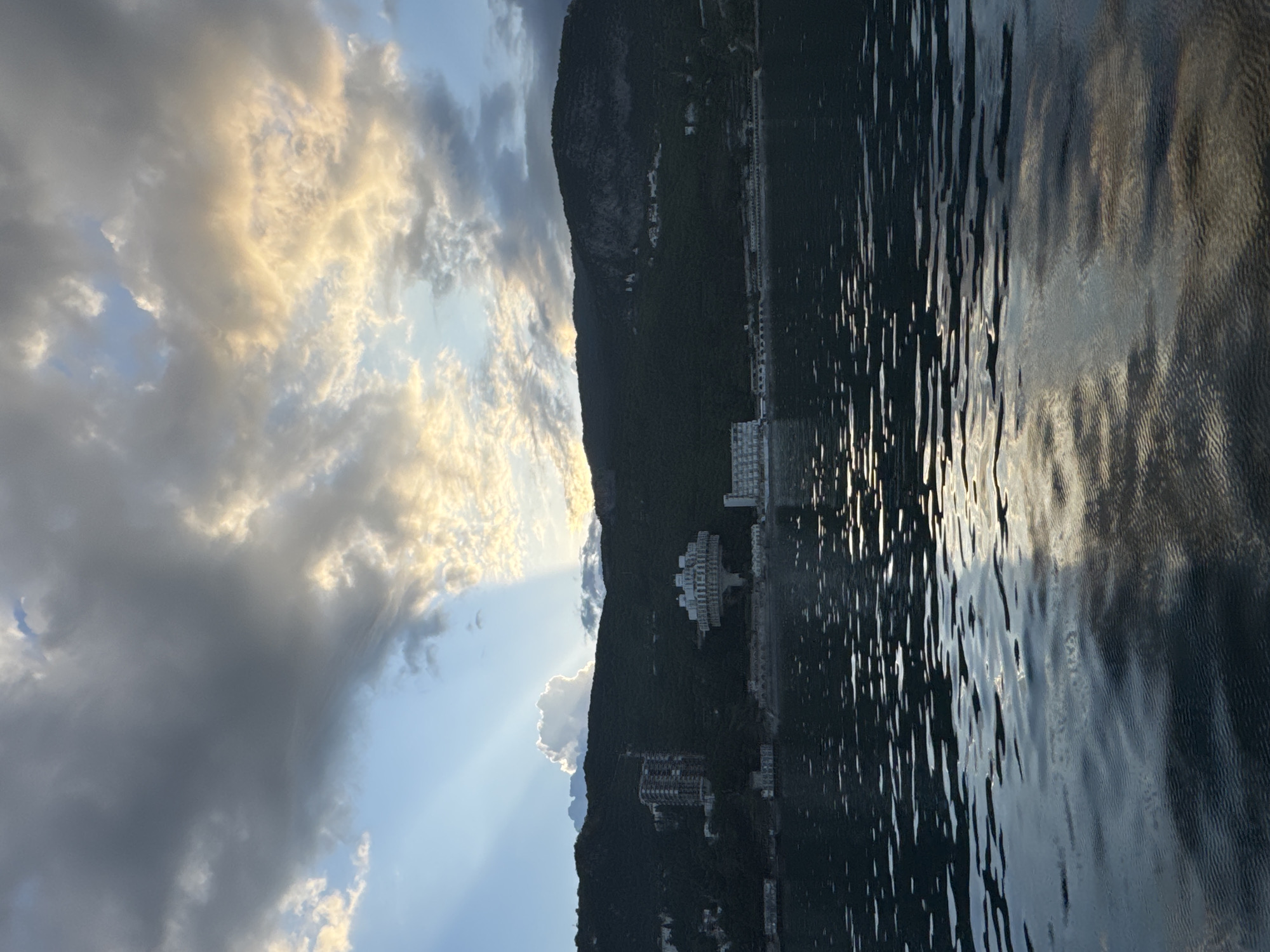 The image captures a shoreline at Гаспринский поселковый совет, Ukraine. A dark, mountainous landscape runs vertically along the right side of the frame, punctuated by buildings. The sky on the left is a blend of clouds and blue sky with sun rays beaming through. The body of water to the right reflects the light and sky.