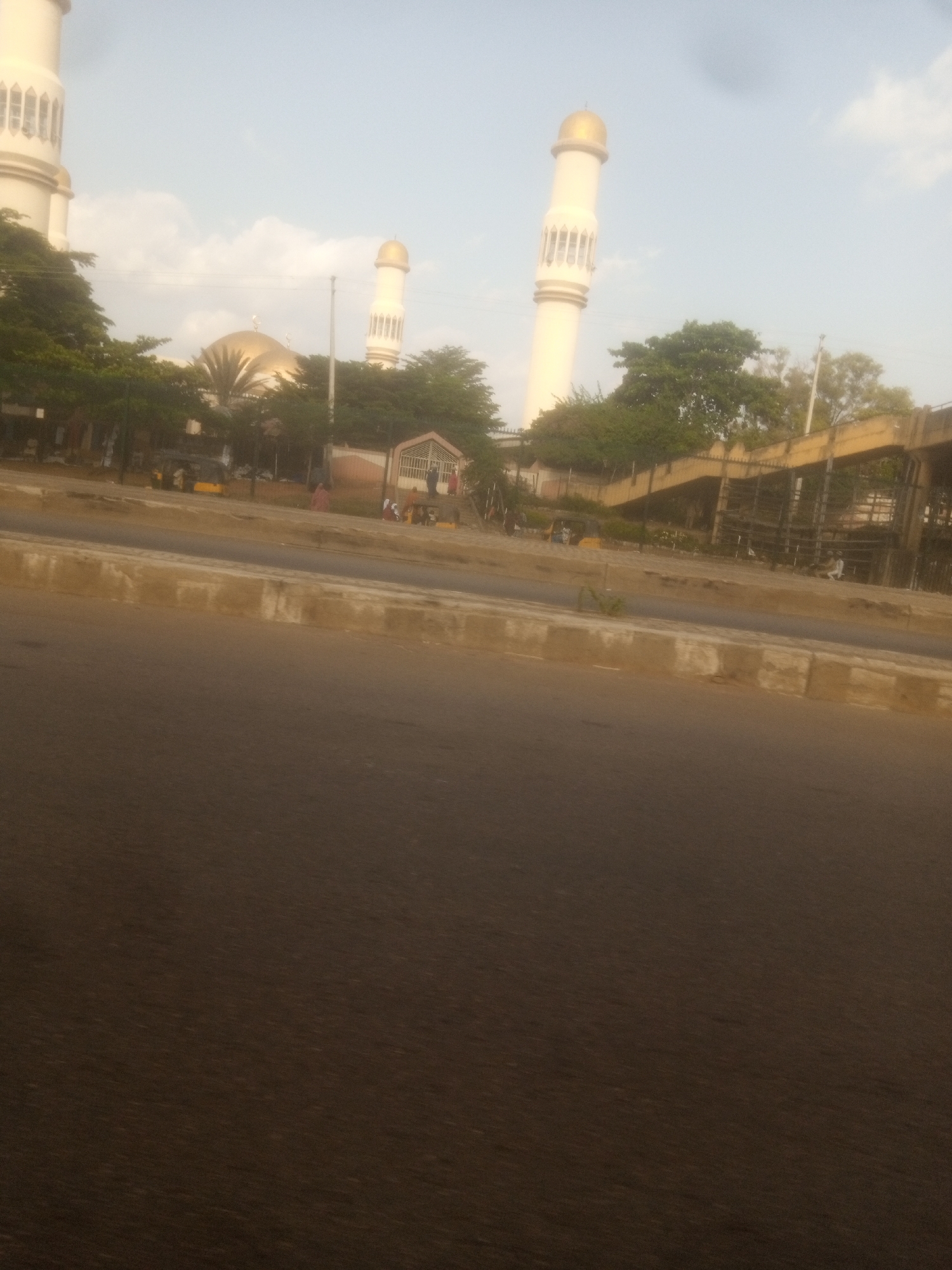 The image captures a street scene in Unguwar Sarki, Nigeria, with a prominent mosque featuring a golden dome and two tall minarets. The mosque is partially obscured by lush green trees. In the foreground, a paved road stretches across the lower portion of the frame, with a concrete divider separating it from a sidewalk area. Behind the divider, a yellow vehicle, possibly a trishaw, is visible, along with several individuals walking or standing near a building with a white gate. Further in the background, an elevated pedestrian walkway or bridge construction is evident. The sky above is a pale blue with scattered clouds, suggesting a daytime setting.