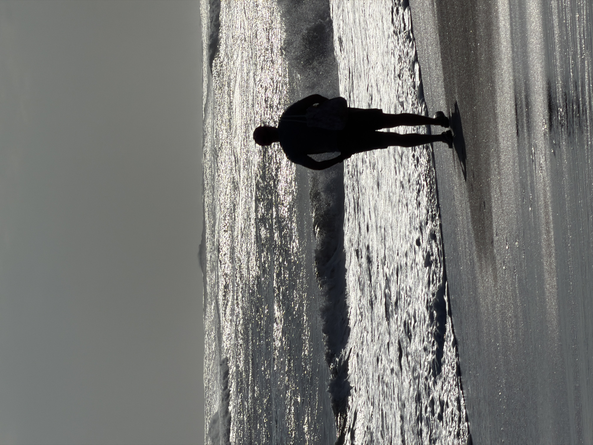 A person, silhouetted against a bright, overcast sky, stands on a dark, wet beach facing the ocean. The person is wearing shorts and a t-shirt, with a bag slung over their shoulder. Waves are breaking on the shore, and the water reflects the sky's light. The scene is devoid of other people or man-made structures, suggesting a secluded location. The overall mood is one of solitude and contemplation. The location is the northern coast of Kuta, Bali, Indonesia, a popular tourist destination known for its beaches and surf breaks.