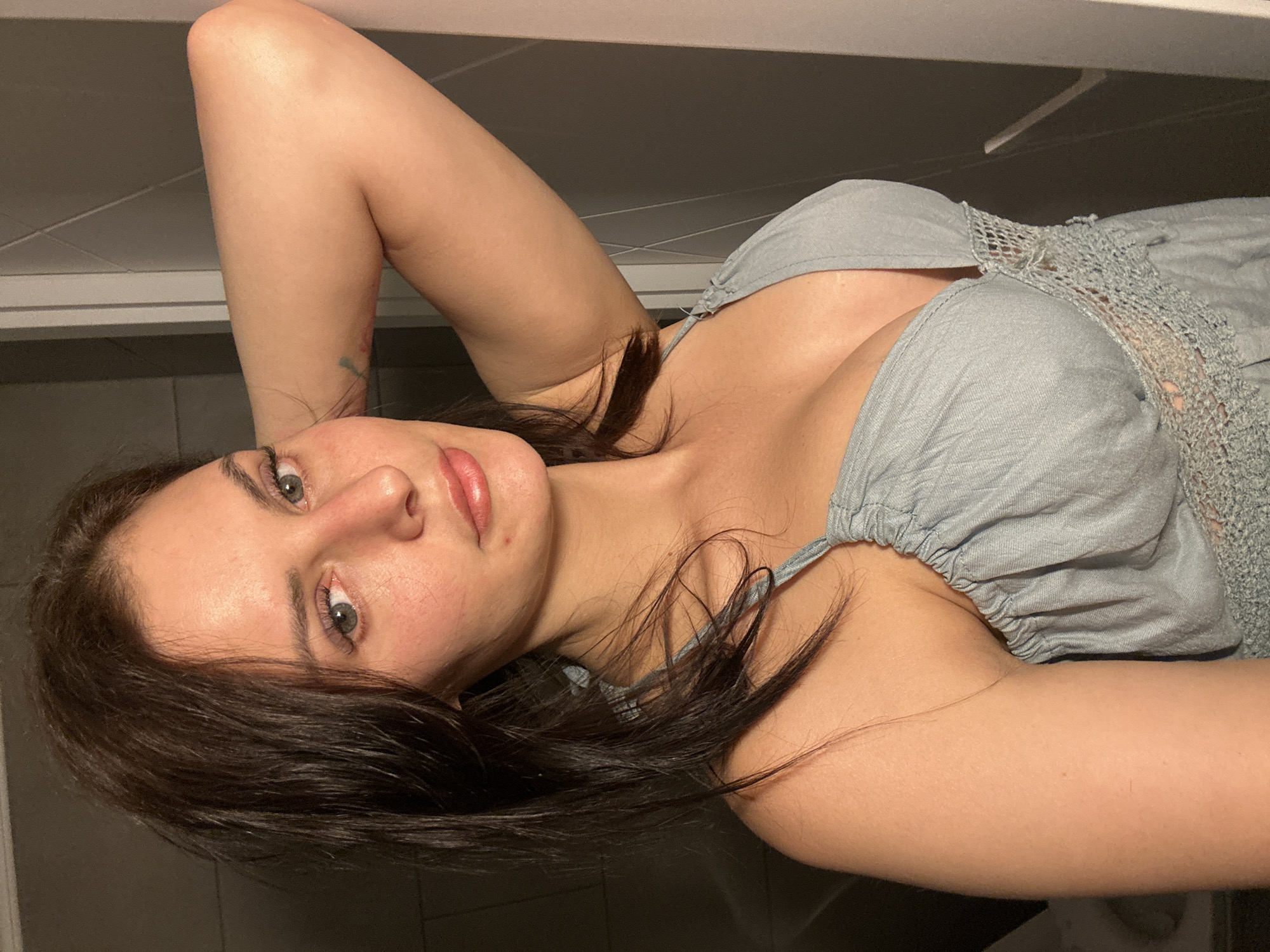 An outdoor, waist-up, eye-level, medium shot captures a fair-skinned woman with dark brown hair and green eyes. Her left arm is bent at the elbow, with her hand resting on the back of her head, exposing her armpit. She is wearing a light blue, sleeveless dress with thin straps and a low-cut neckline adorned with lace trim. Her lips are slightly parted, and she has a subtle smile. The background consists of a dark, tiled wall on the left and a lighter, textured wall on the right, with a vertical white element suggesting a door frame or partition. The lighting is dim, with shadows visible.