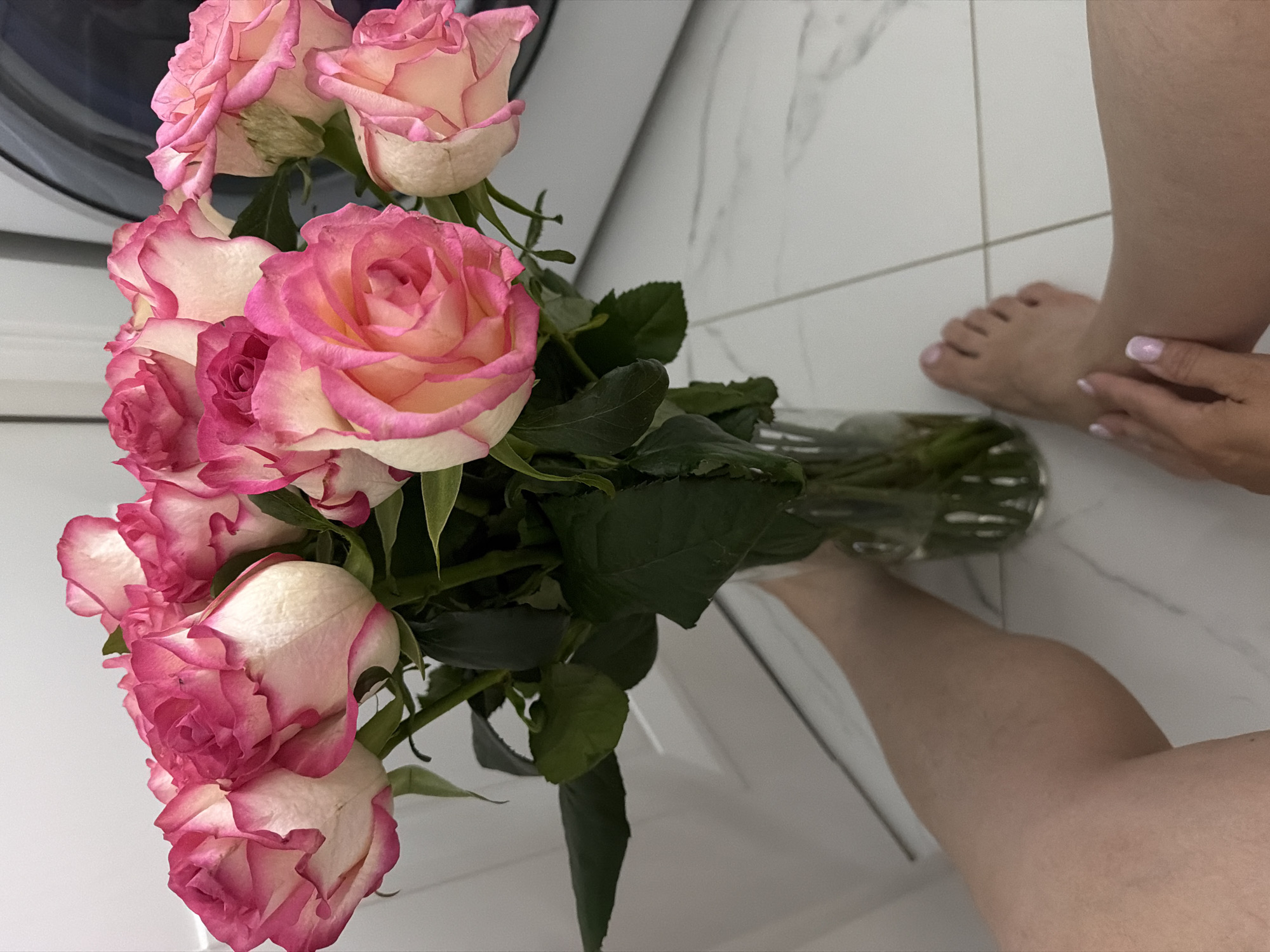 A still life photograph, seemingly taken from above, shows a bouquet of pink and white roses in a clear glass vase on a white tile floor. A person's legs and feet are also visible in the frame. The location is suggested to be the city of Votkinsk, Russia. The environment appears to be a bathroom, indicated by the tile floor and a white washing machine on the left. The person's hand is near their foot with manicured nails. The roses are the primary subject.