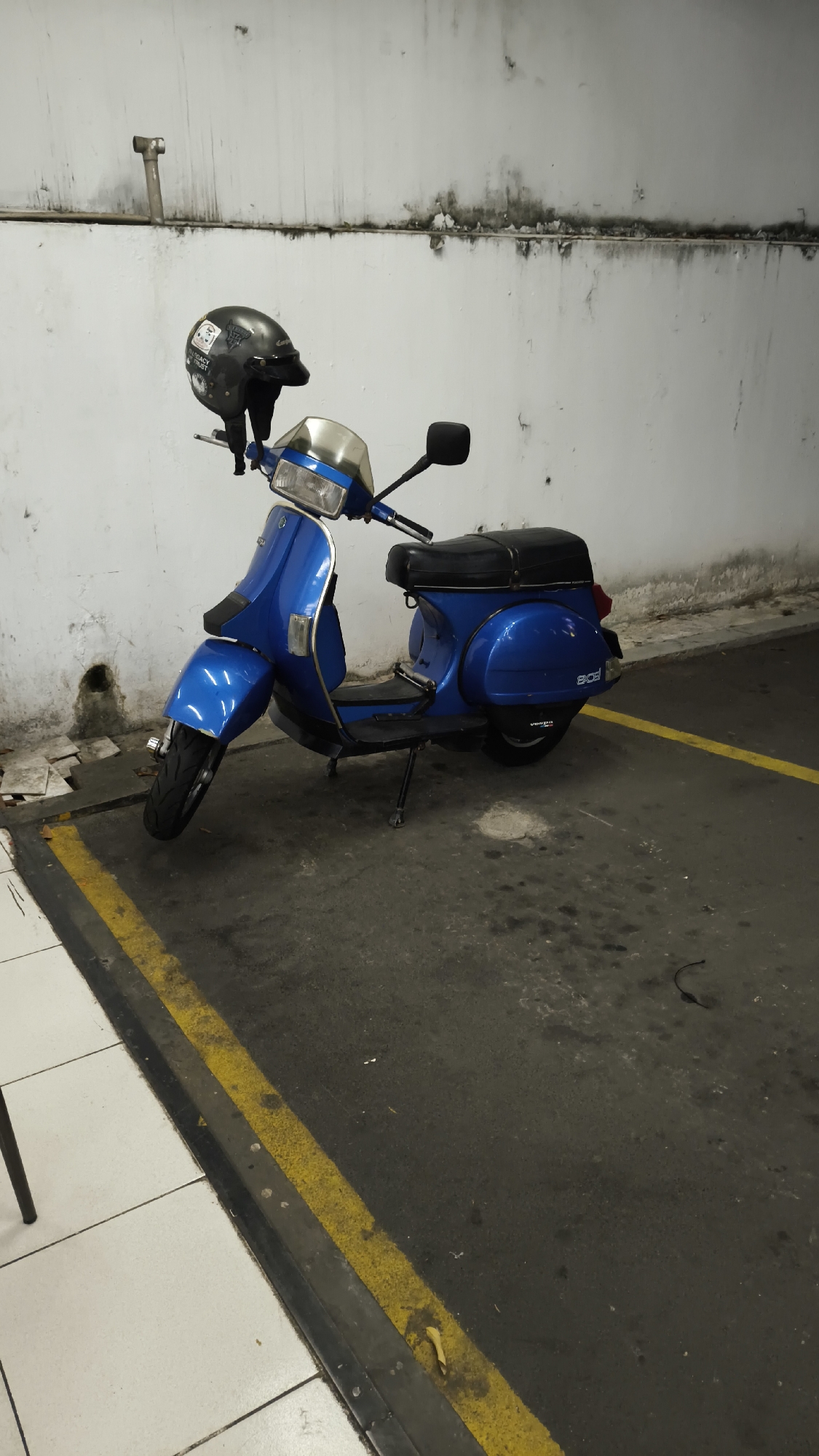 In South Jakarta, Indonesia, a blue Vespa scooter is parked on an asphalt surface, distinguished by yellow and black painted lines. The scooter's handlebars are adorned with a black helmet featuring a white graphic. The background is a textured, off-white wall showing signs of dampness and dark streaks. To the left of the scooter, white tiled flooring is visible, separated from the asphalt by a dark border. The scene is illuminated by artificial light, casting subtle shadows. No individuals are present in the frame.