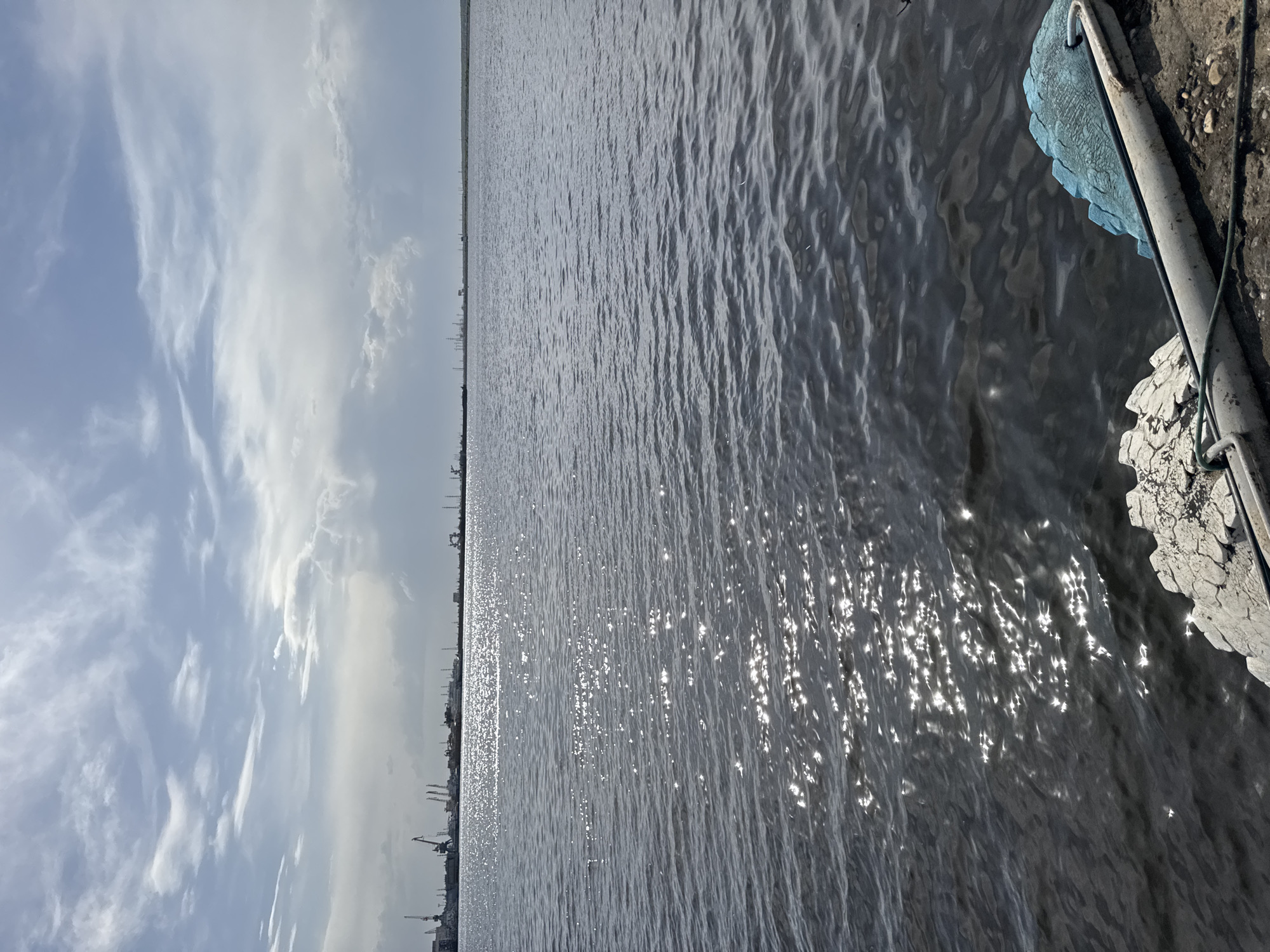 The image shows a body of water reflecting sunlight, with a clear sky and clouds visible in the background. On the right side of the frame, part of a dock or structure is visible, with a blue and white bumper or fender attached. In the distance, a skyline is discernible, potentially indicating an urban or industrial area. Considering the prompt indicates the location is Chaikovsky, Russia, the skyline could be related to industrial facilities or the city itself.